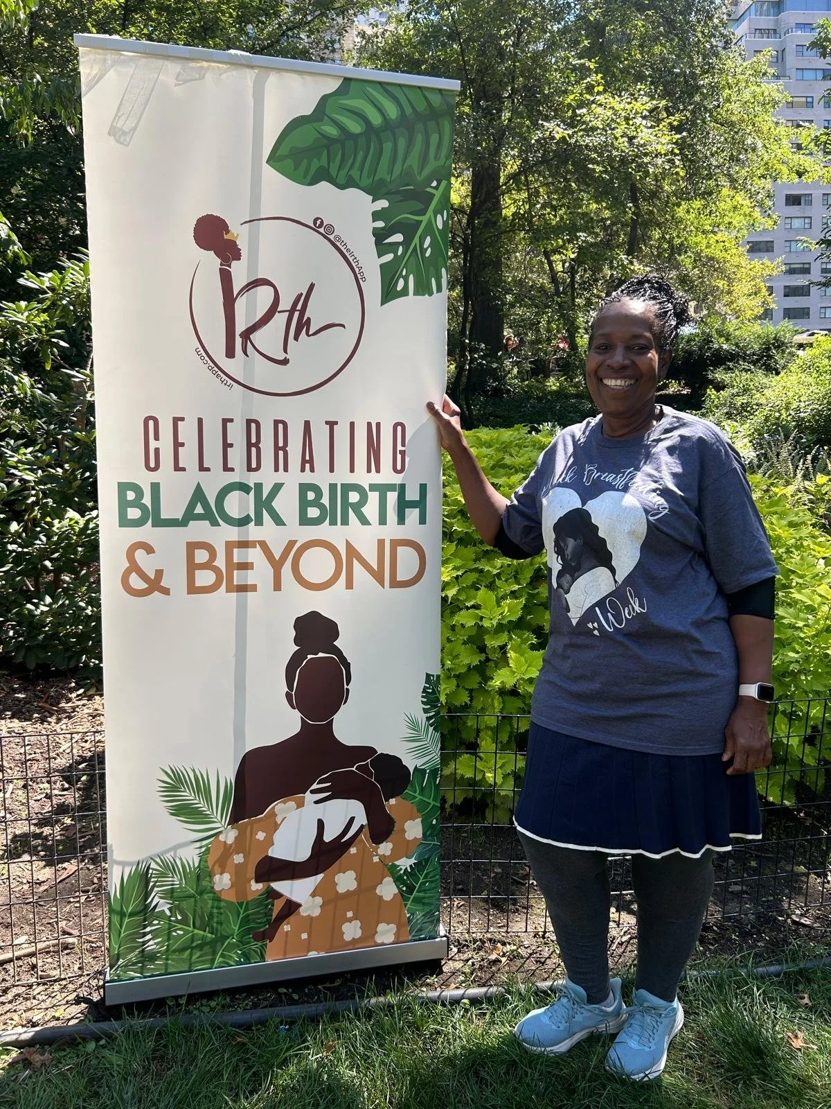 Dawnette Clarek-Anglin in a blue T-shirt and black skirt, smiling and standing outdoors next to a colorful banner that says 'Celebrating Black Birth & Beyond'