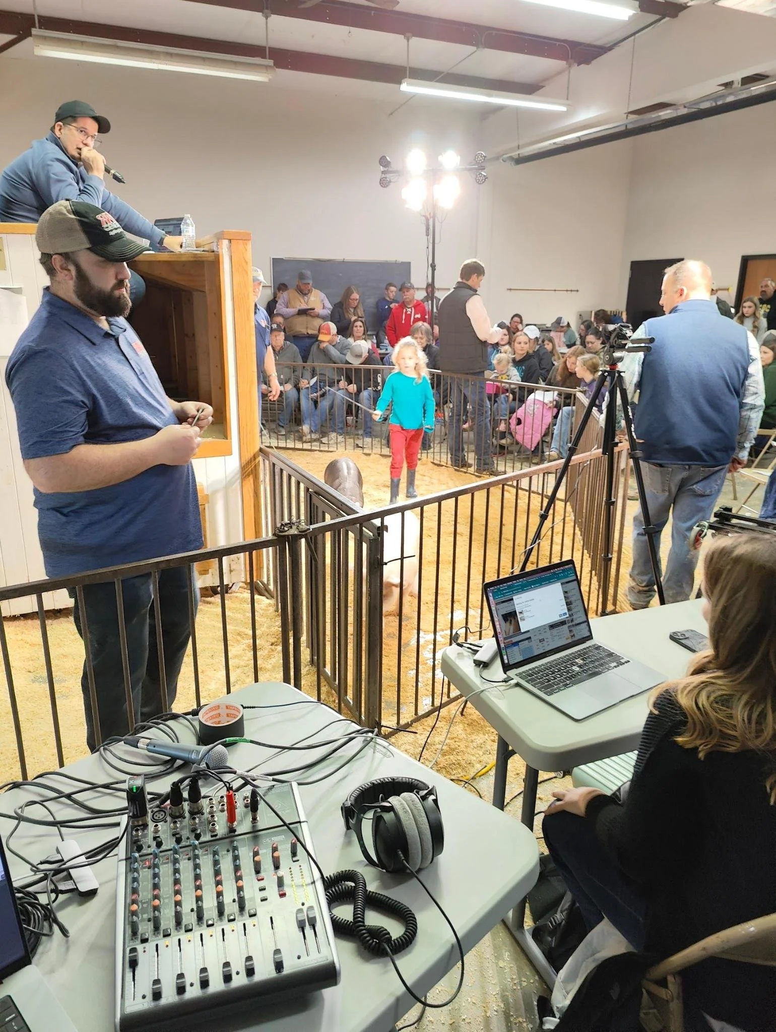 A live event or auction is happening in a large room filled with people. There are individuals at a desk with audio equipment, including a mixer and headphones, suggesting a sound or broadcasting setup. Bright lights are illuminating the scene. The a