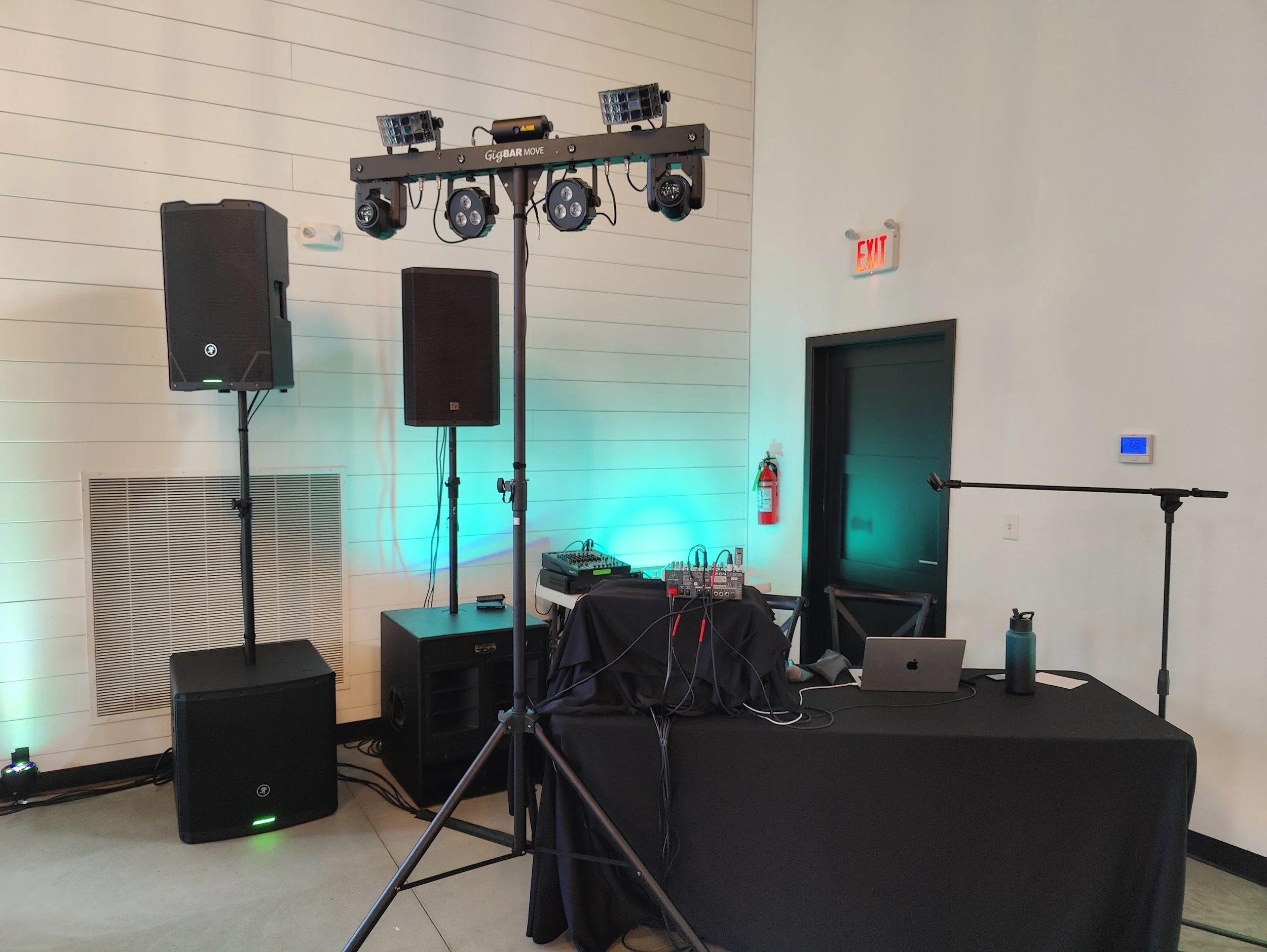 Audio and stage lighting equipment setup on a black stage, including speakers, a mixer, microphones, and lighting fixtures, in a room with white-paneled walls, an exit sign, and a door.