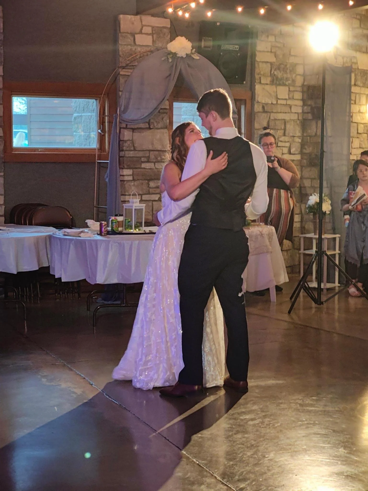 A couple dancing closely at a wedding reception, with a woman in a striped dress and glasses taking photos in the background.