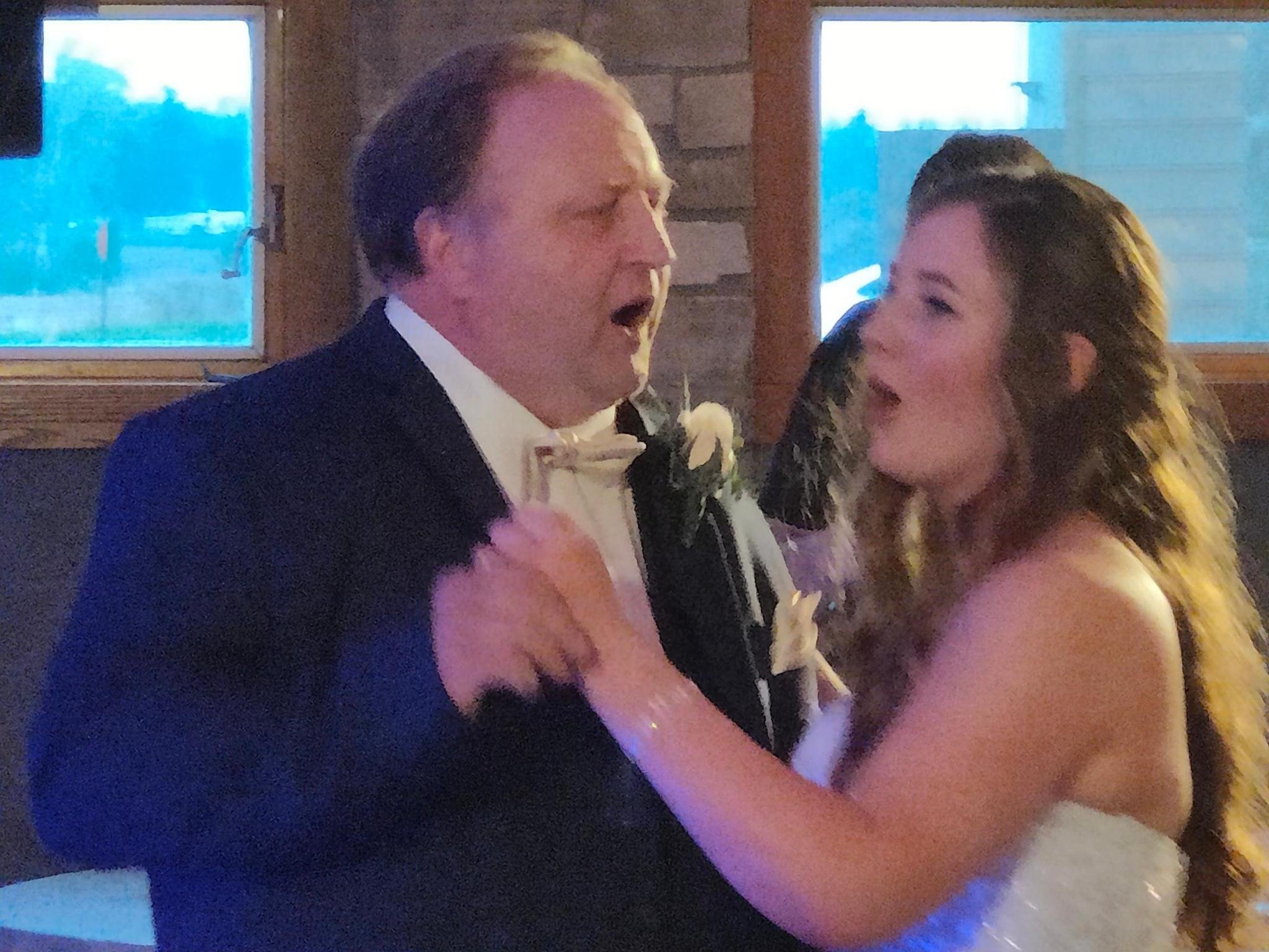 A man and a woman are dancing together indoors, singing or talking with smiles. The man is wearing a tuxedo with a bow tie, and the woman has curly hair and is wearing a sleeveless dress.