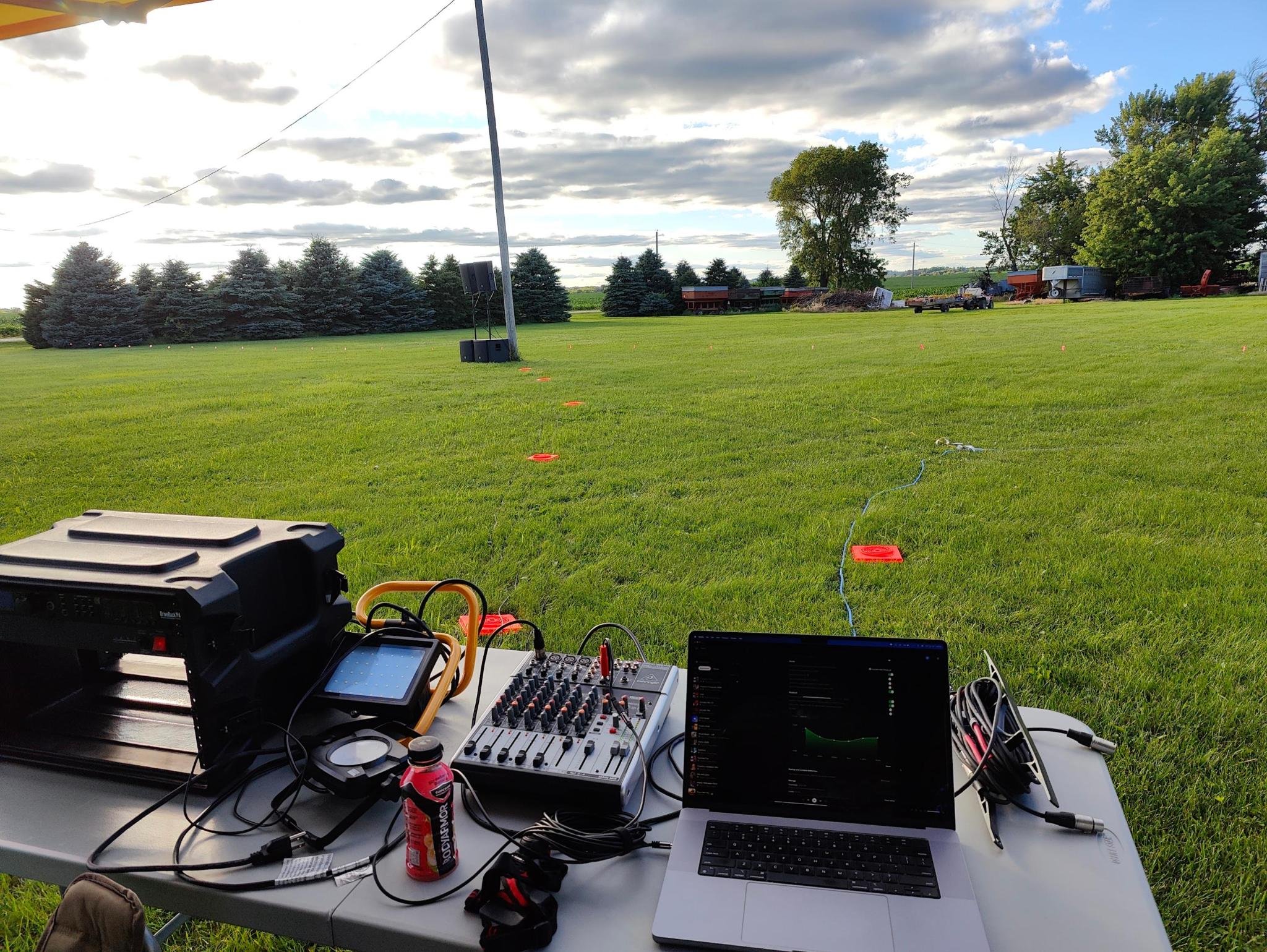 Audio equipment setup on a white table outdoors with green grass, trees, and cloudy sky in the background. The setup includes speakers, wires, laptop, audio mixer, and other electronic devices.