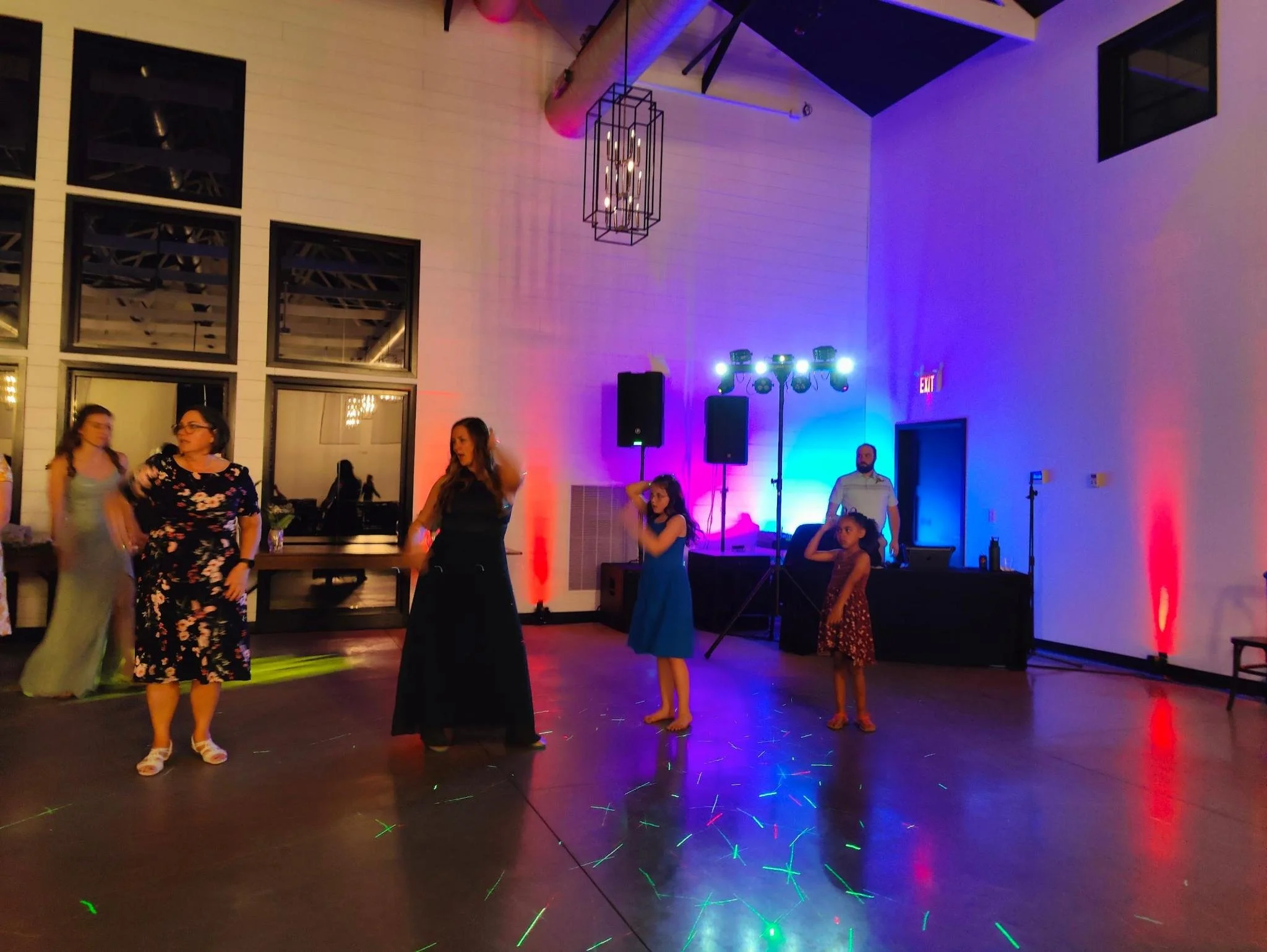 People dancing and socializing in a large indoor event space with colorful lighting, a high ceiling, and wall windows.
