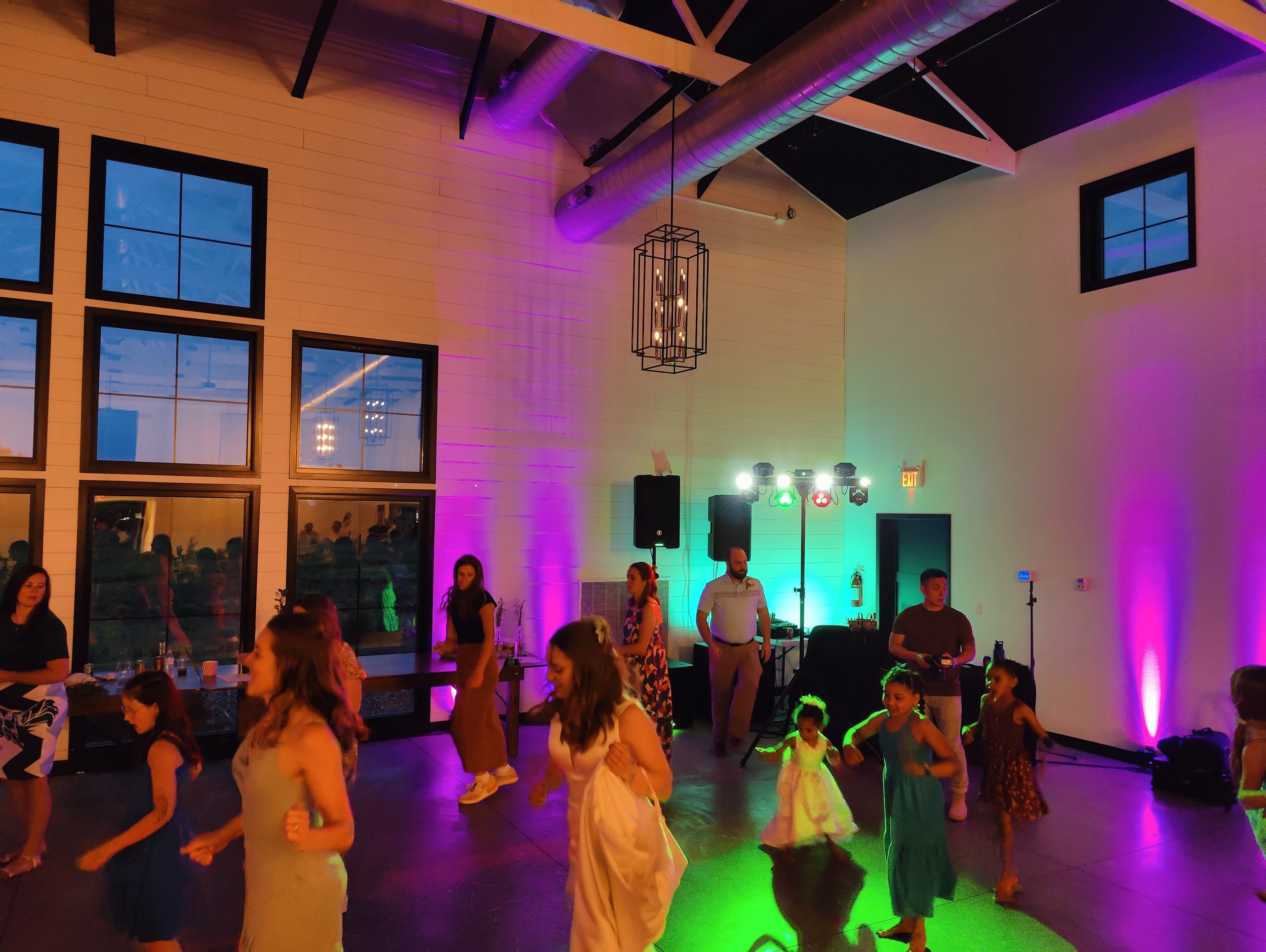 People dancing at a party or celebration in a large indoor space with colorful lighting and windows showing an evening sky.