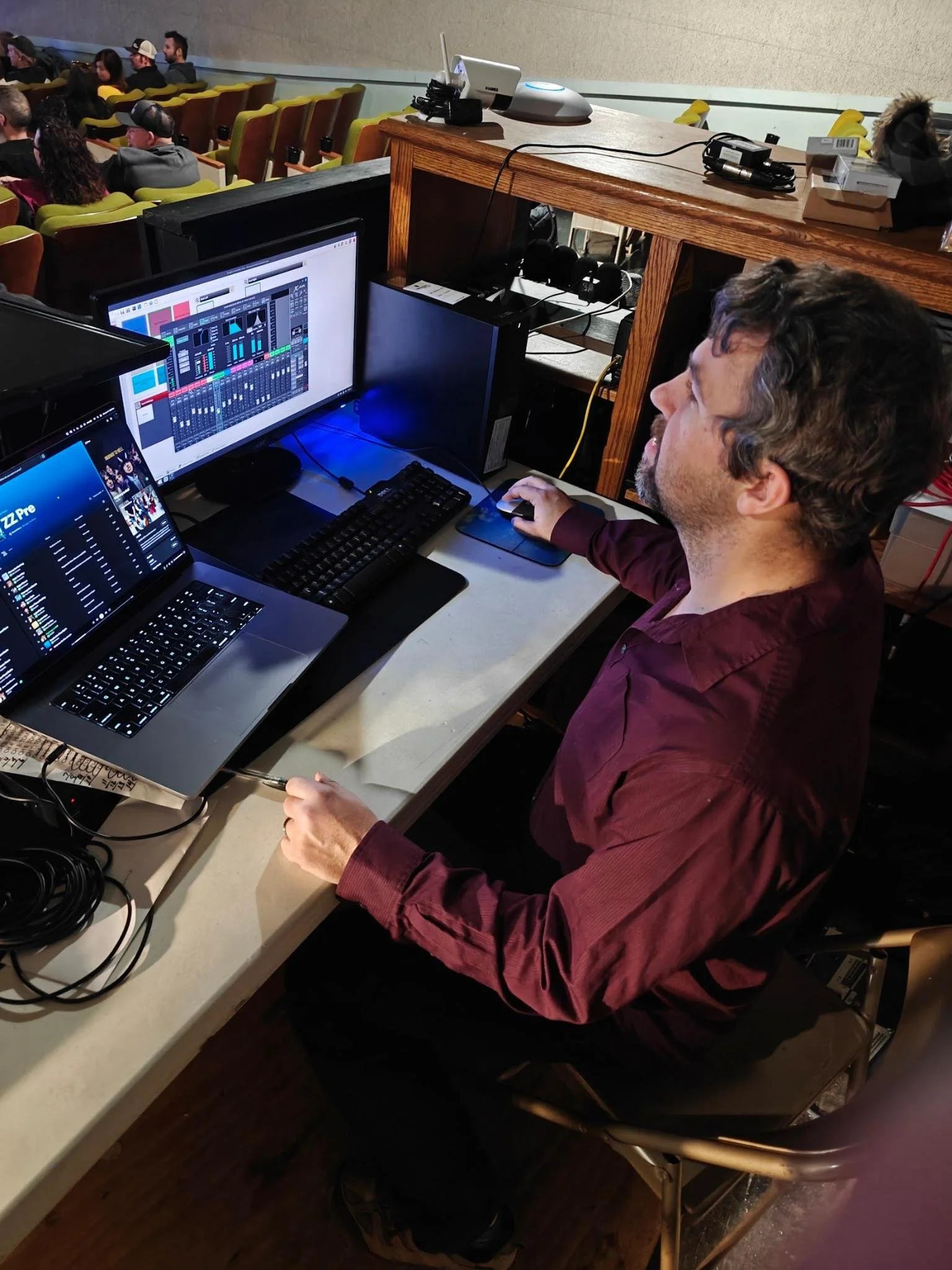 A man with curly hair and a beard wearing a maroon shirt sitting at a sound engineer's station with multiple monitors, a keyboard, and a mouse, working on audio mixing during a concert or event, with audience seated in the background.