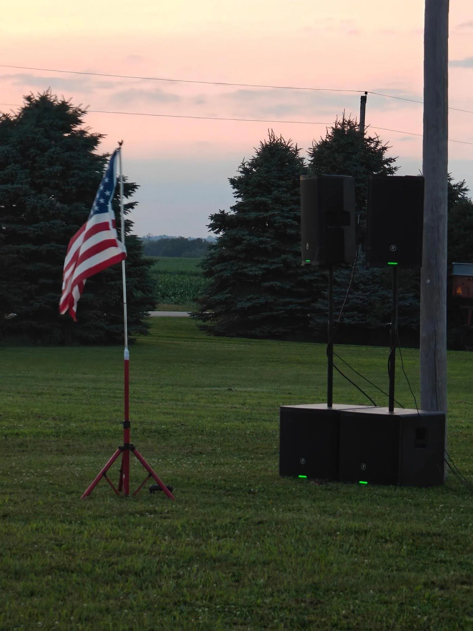 An outdoor scene at dusk with a pink and blue sky, a row of evergreen trees, a flagpole with the American flag, and speakers with green lights.
