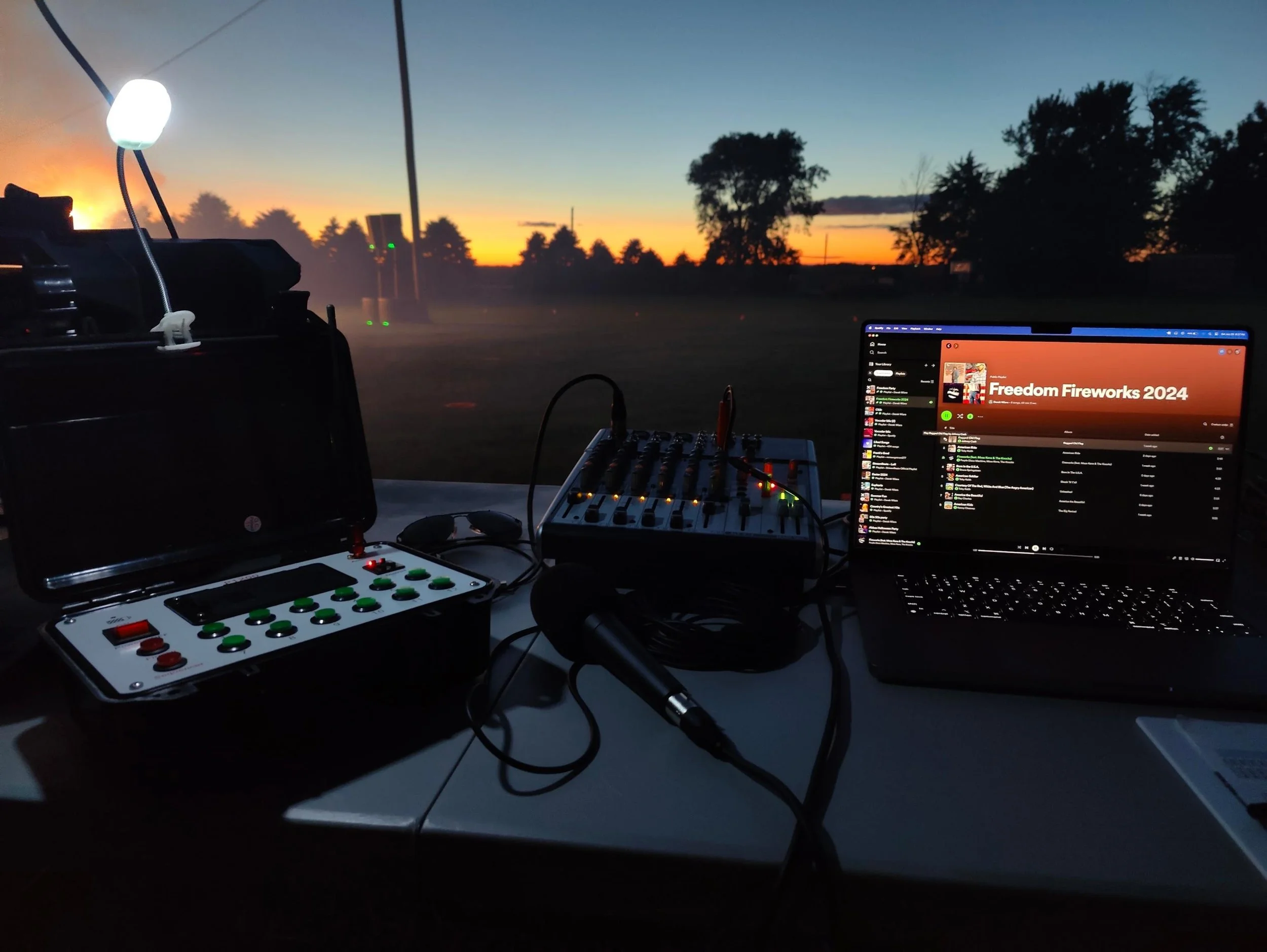DJ equipment setup with a laptop displaying 'Freedom Fireworks 2024', a controller with green and red buttons, a speaker, and a light, with a sunset and trees in the background.