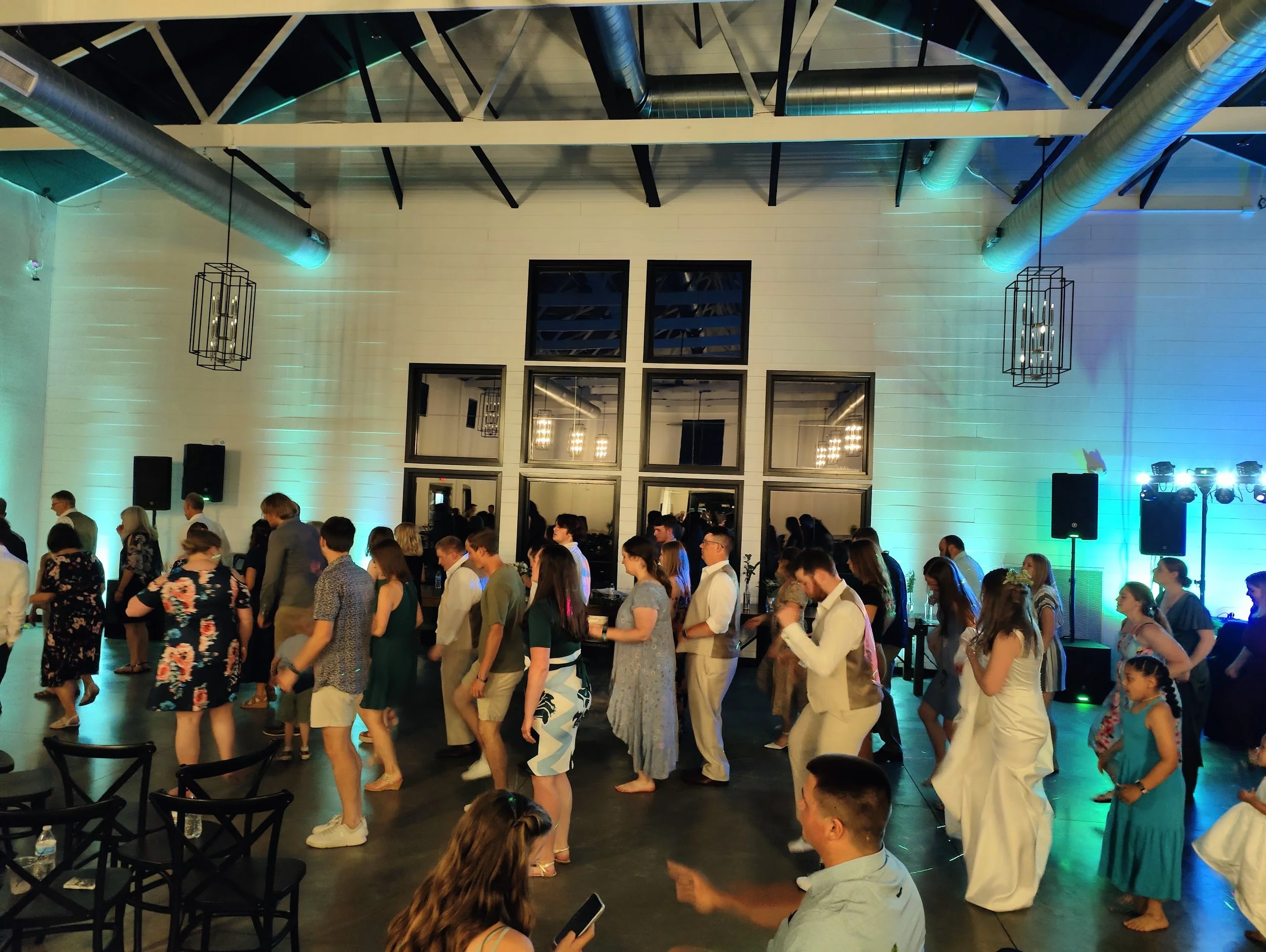People dancing at a social event in a large, modern venue with high ceilings, exposed ductwork, and colorful lighting.