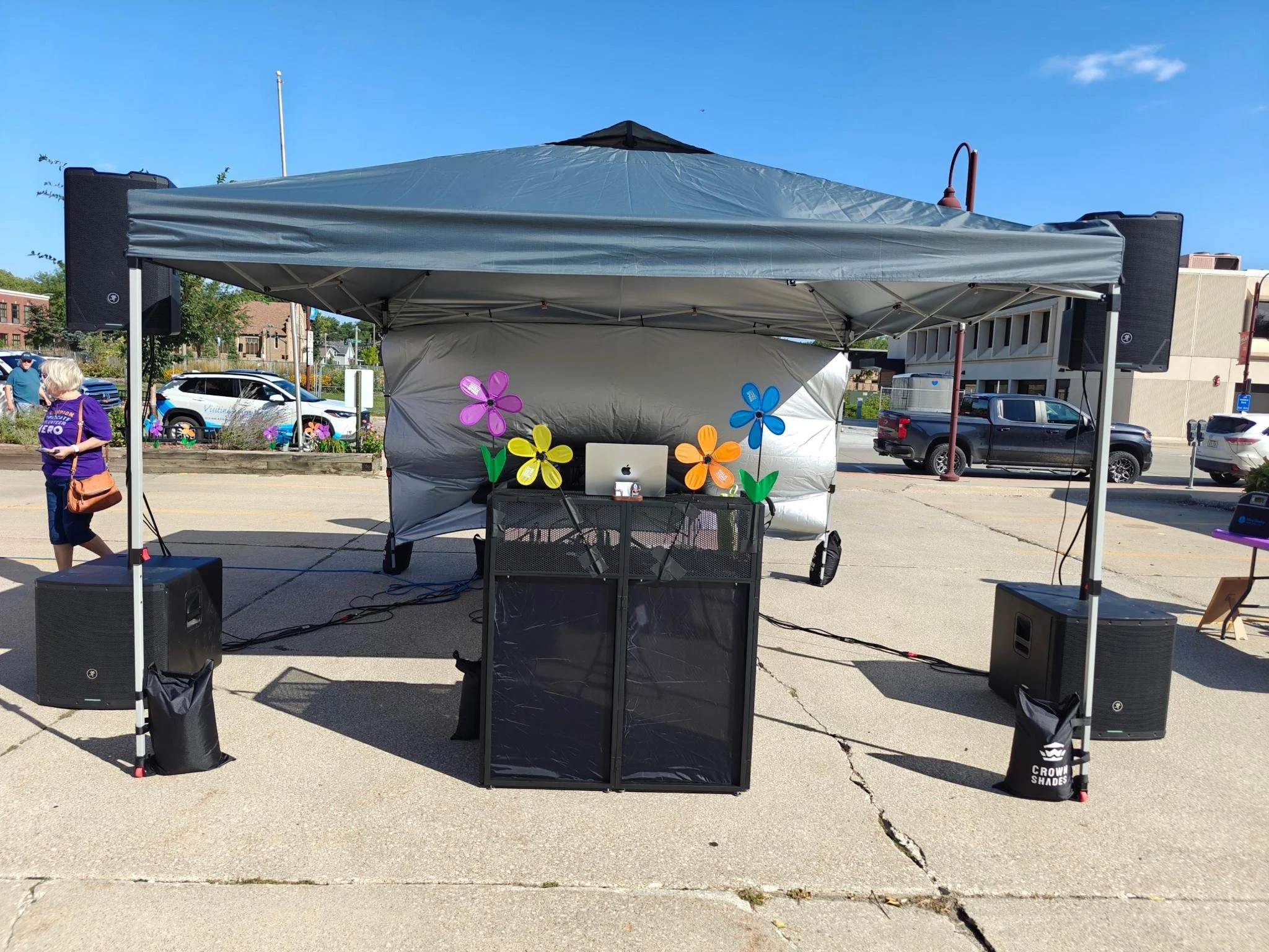 A music stage setup with large speakers, a tent overhead, colorful paper flower decorations, possibly at an outdoor event or festival on a sunny day.