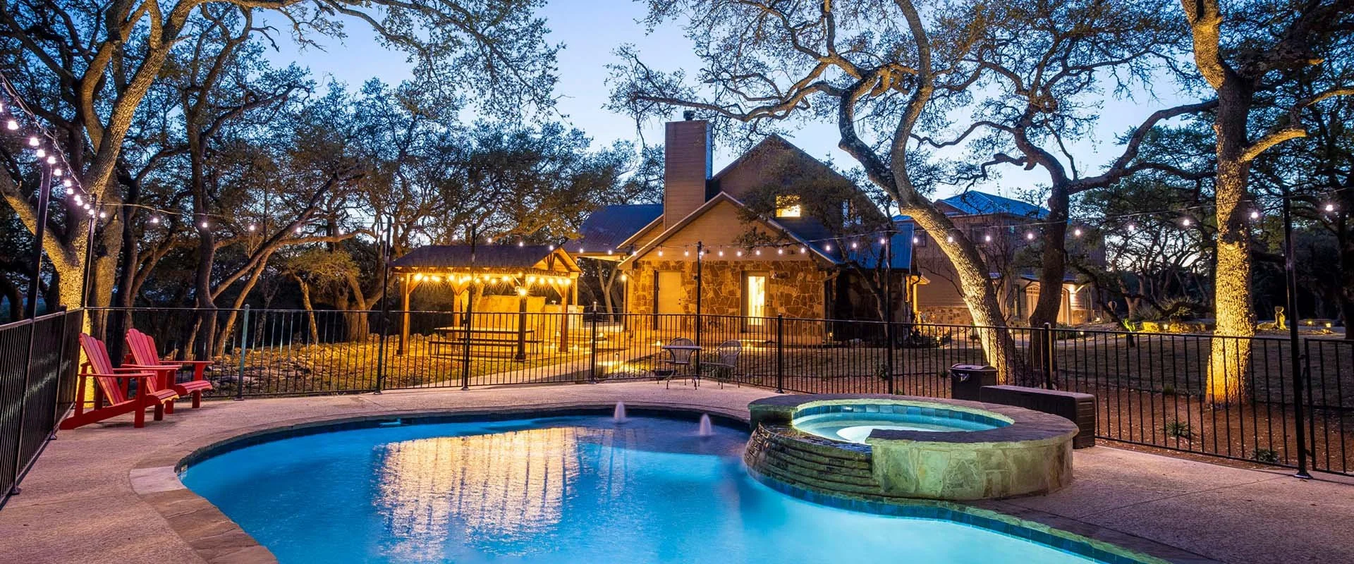 A backyard swimming pool and hot tub are surrounded by a black metal fence. There are red Adirondack chairs near the pool. String lights are hung across the yard, illuminating the trees and a house with a stone facade and a chimney in the background. It is evening or twilight.