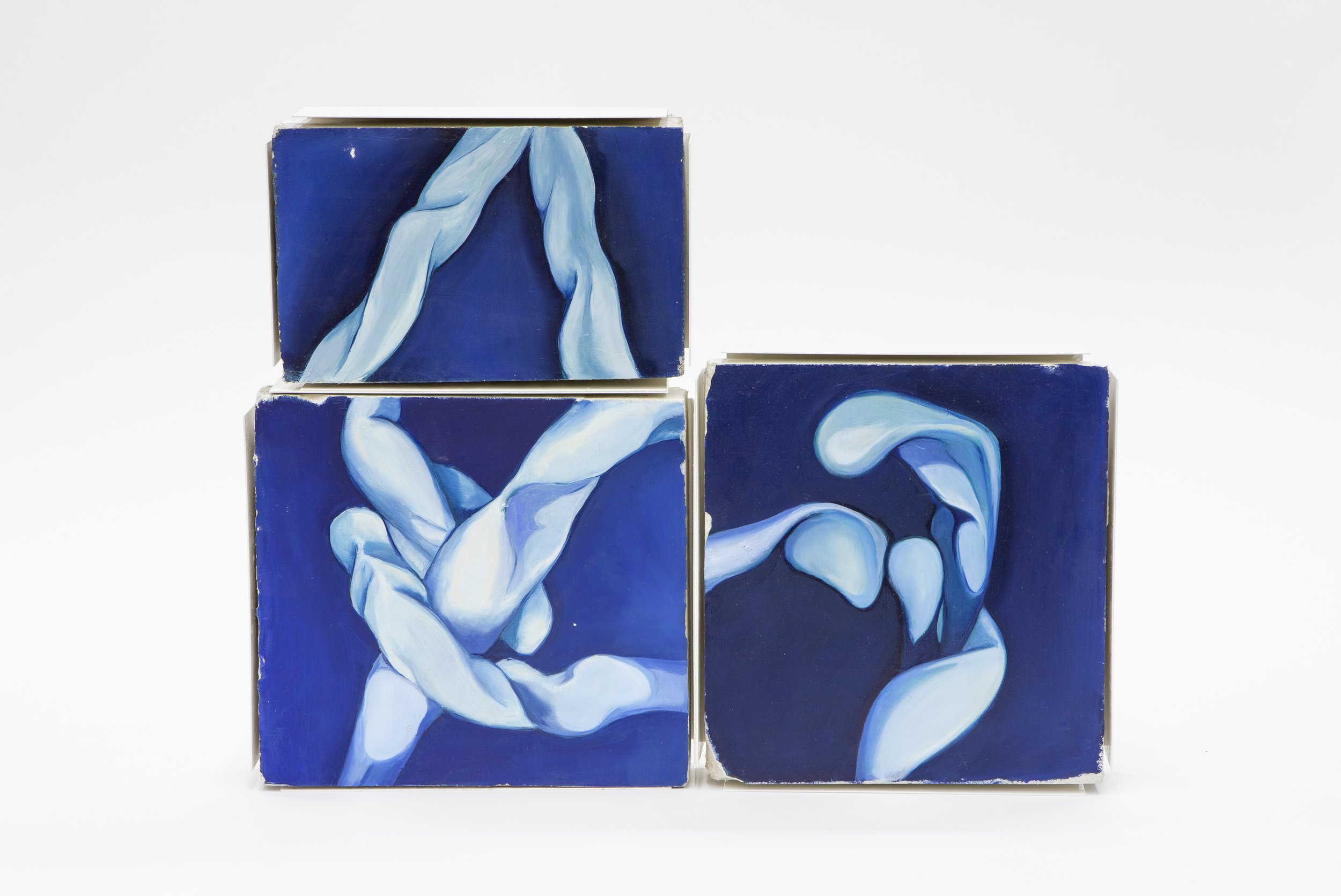 Tres bloques de madera decorados con pintura que representa figuras humanas en tonos azules y blancos, dispuestos en fila sobre fondo blanco.