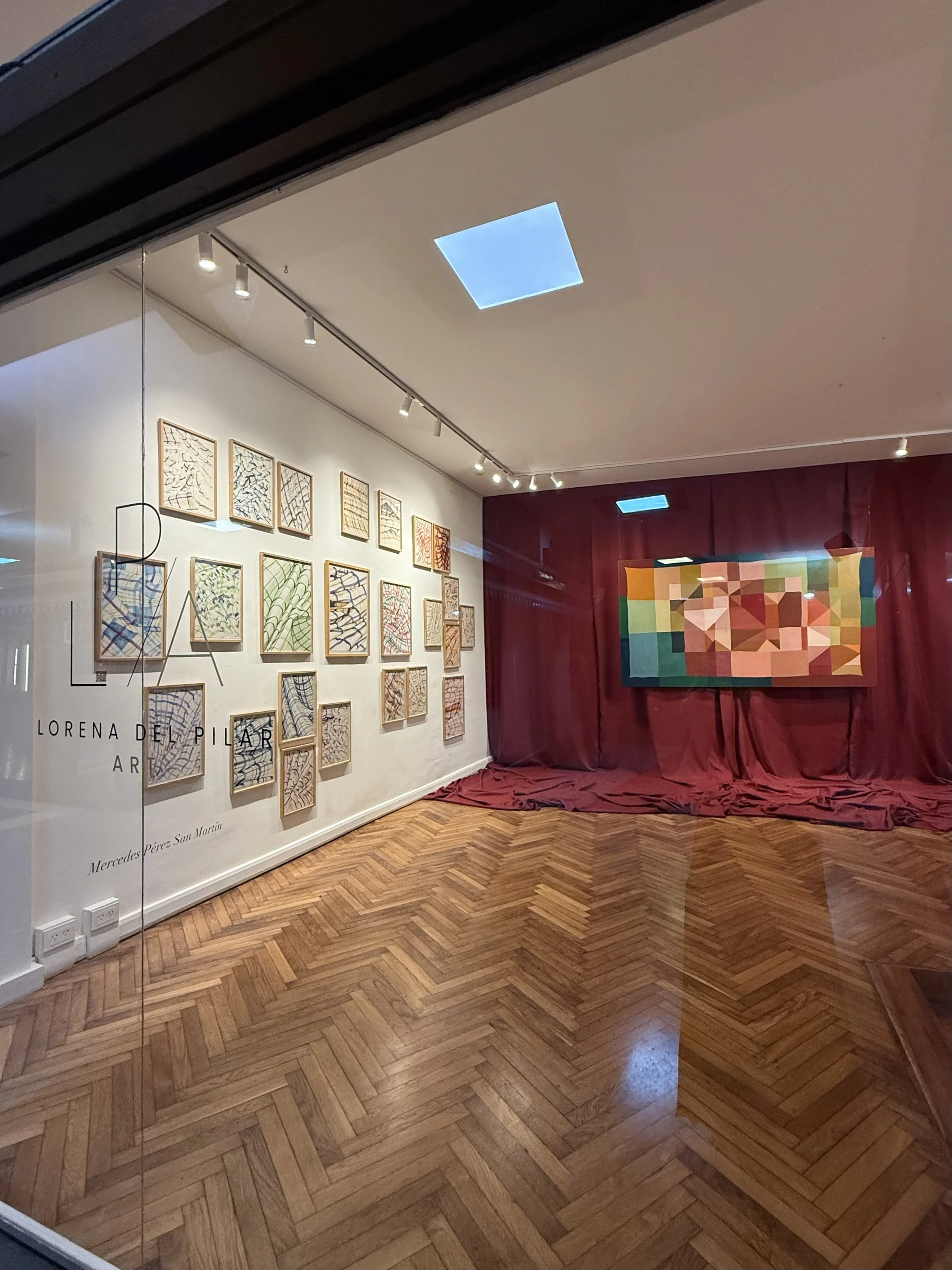 Galería de arte con varias obras en las paredes y un piso de madera; una obra grande con formas geométricas y varios cuadros pequeños con diseños variados, en un espacio con cortinas rojas y plafones con luces
