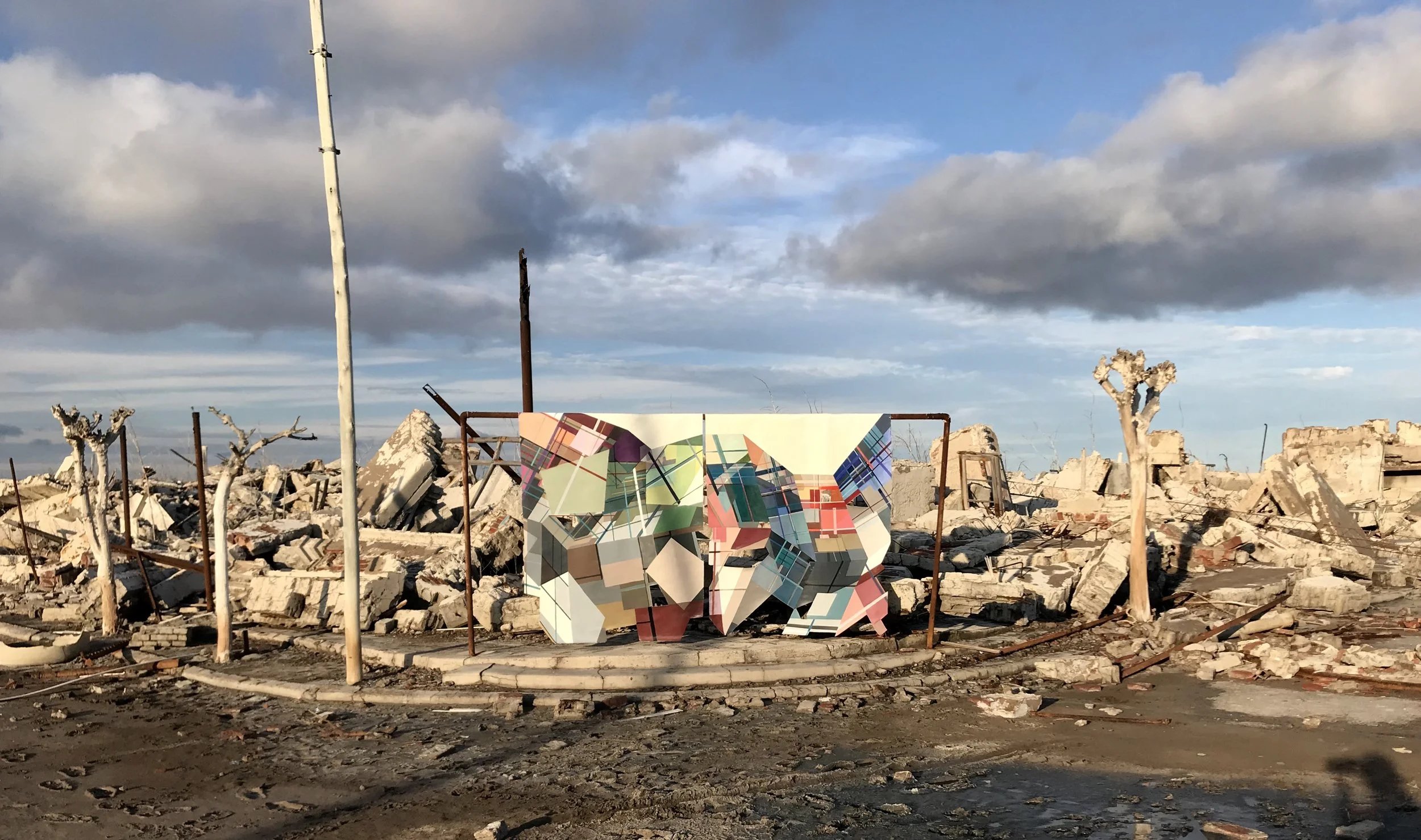Paisaje de escombros de un edificio destruido con un mural colorido en forma de elefante, cielo con nubes grises y azul, en un entorno desolado.