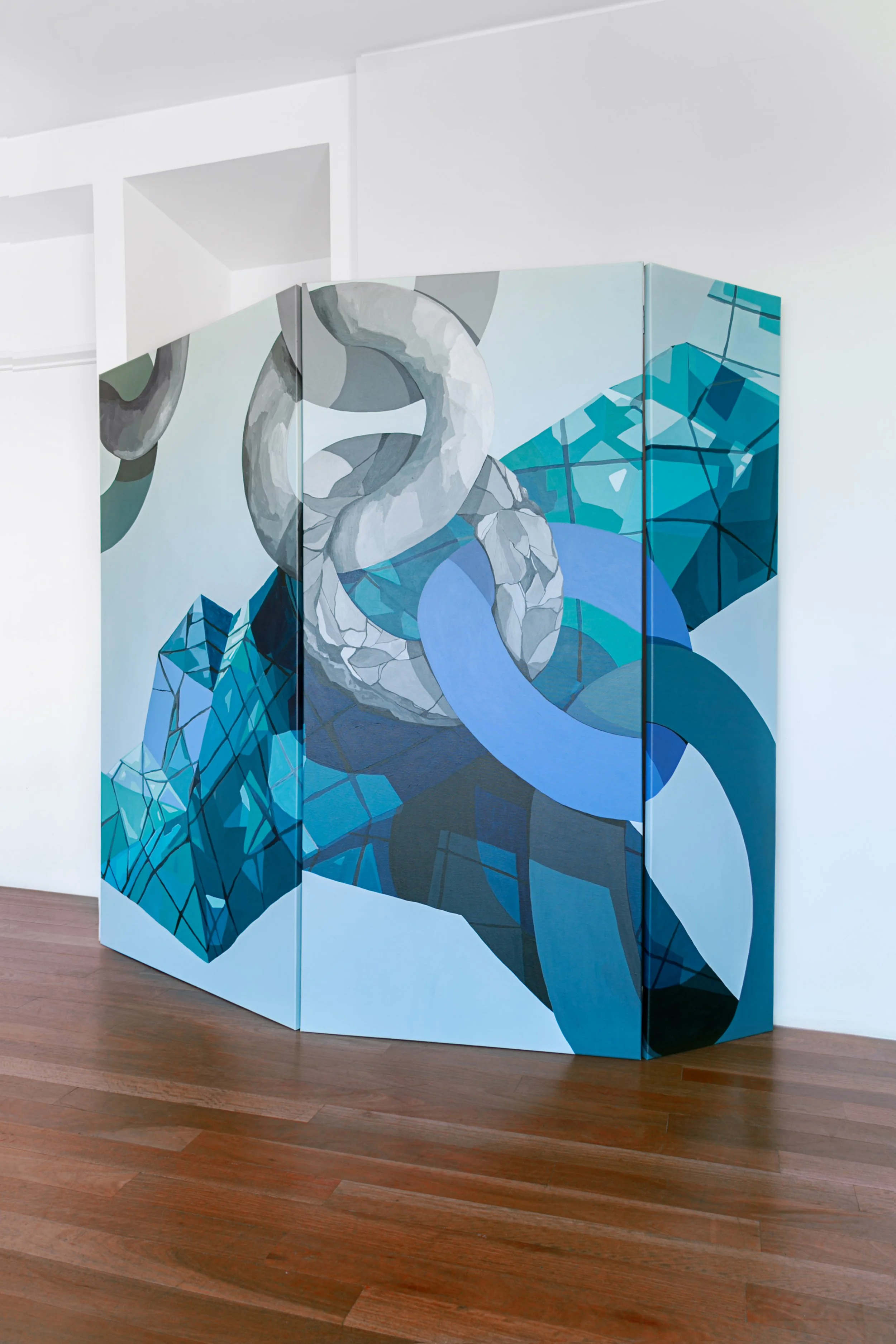 Escultura de arte con formas geométricas en tonos azules, grises y blancos, ubicada en una sala blanca y piso de madera.