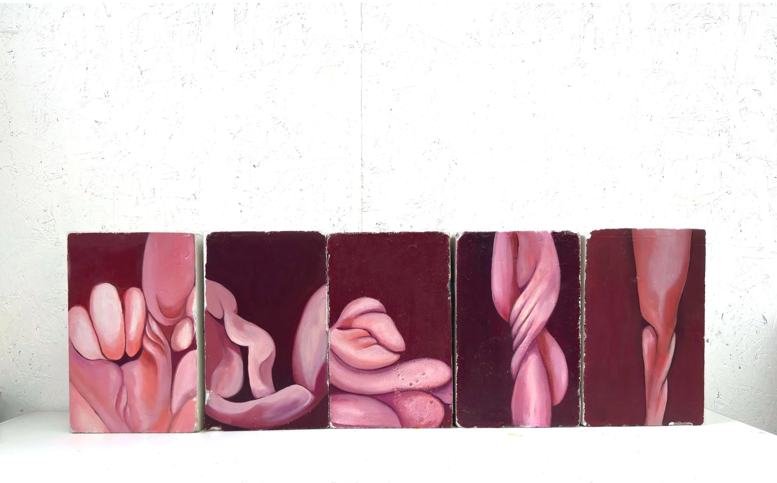 Una escultura abstracta compuesta por seis bloques apilados con formas orgánicas y curvas en tonos rosados y morados, sobre un fondo de pared blanca con textura.