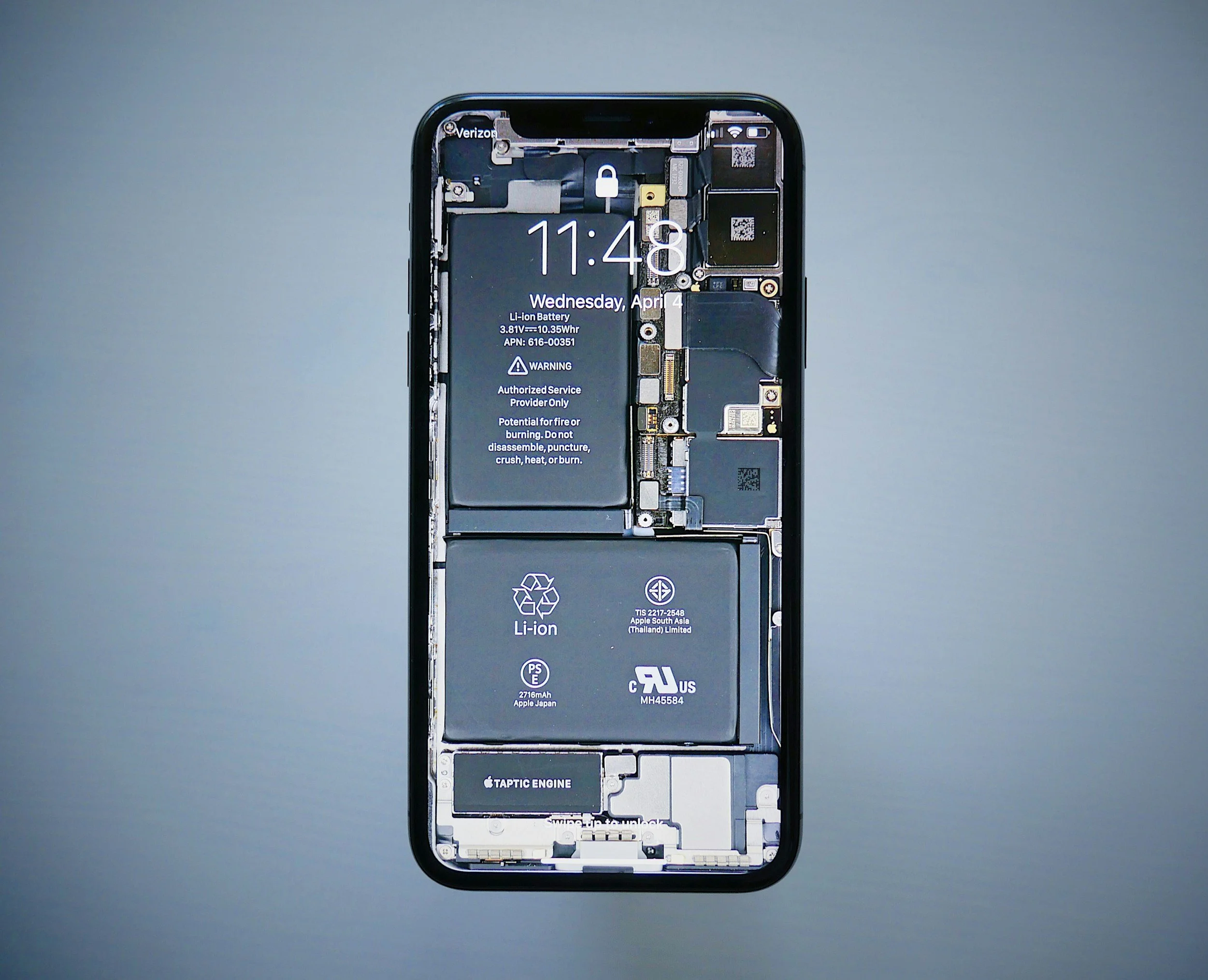 Close-up of a disassembled smartphone showing internal components including a battery, circuits, and connectors, with the screen displaying the time, date, and various warning and information labels.