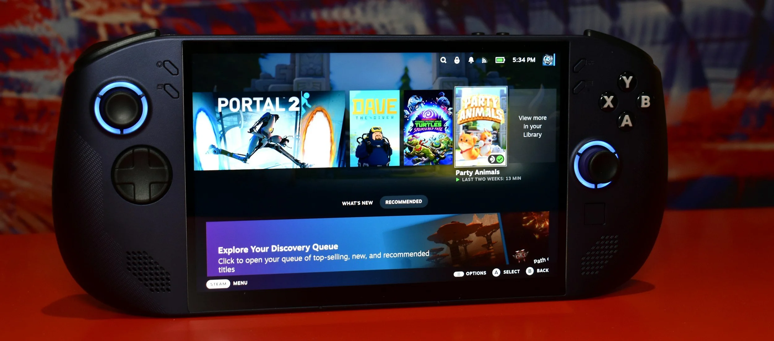 A handheld gaming device displaying a game library including titles like Portal 2, Dave the Diver, Teenage Mutant Ninja Turtles, and Party Animals, with a red surface and a patterned background.