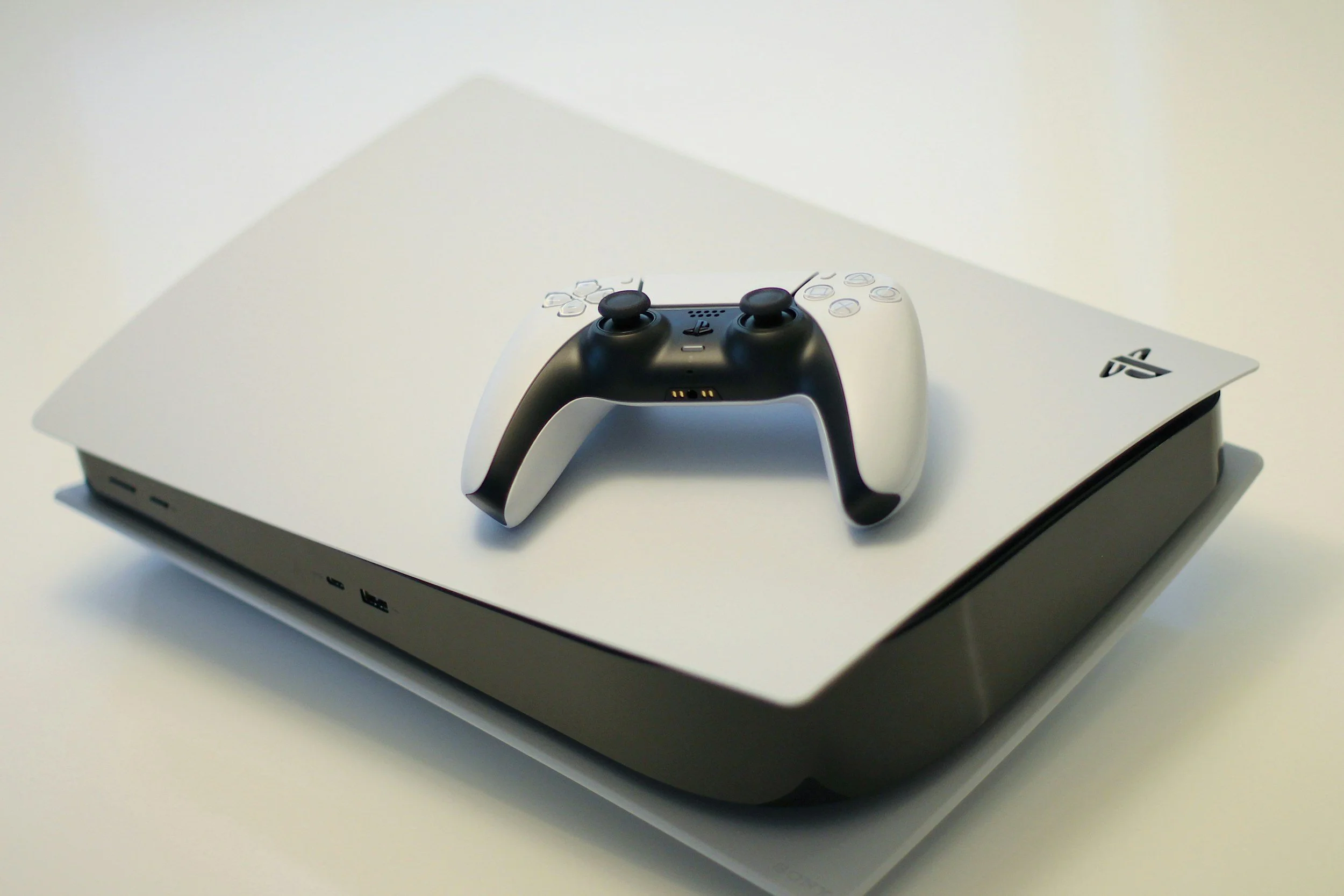 A PlayStation 5 console with a black and white DualSense controller resting on top, on a white surface against a plain white background.