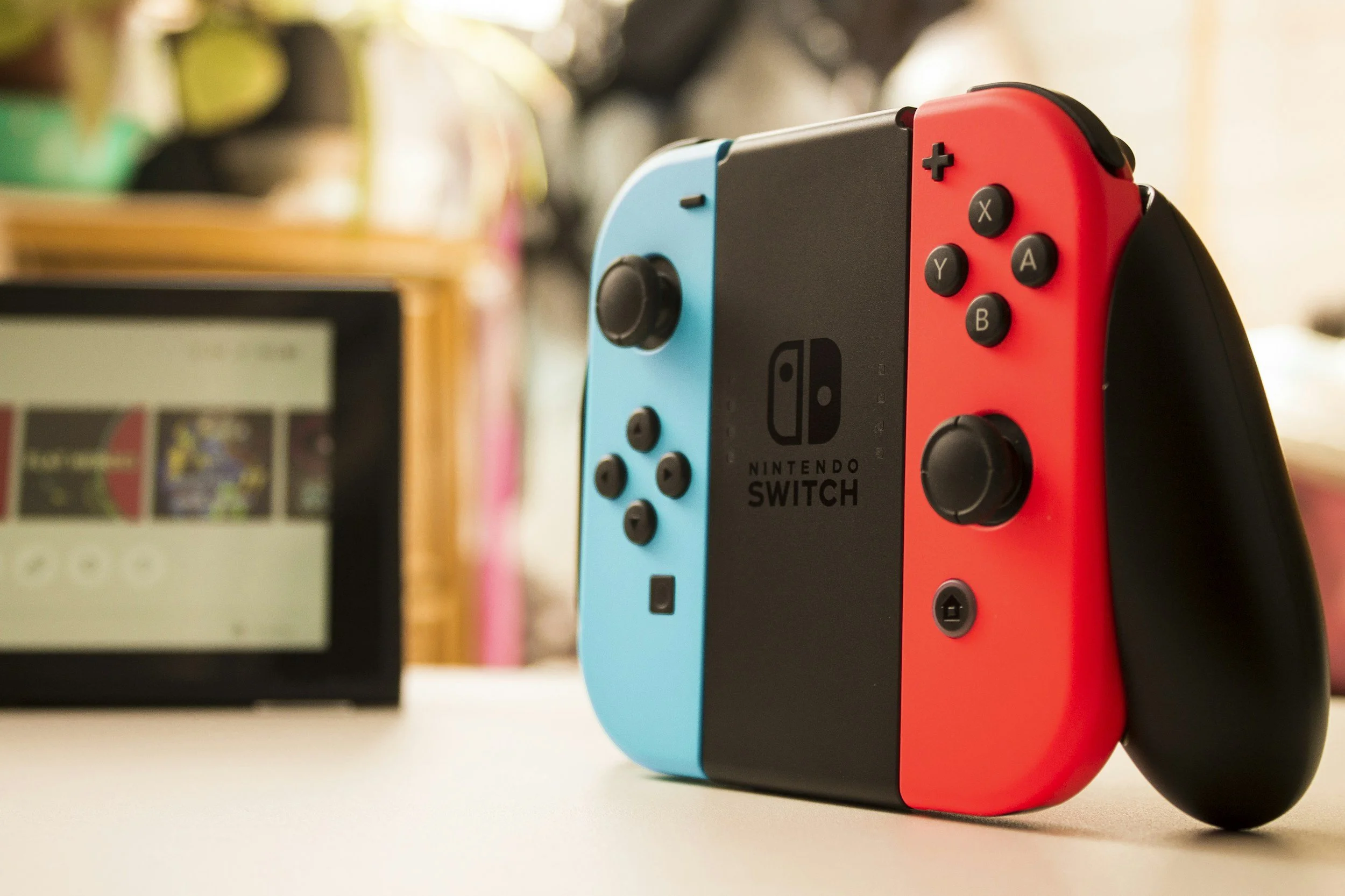 A Nintendo Switch gaming console with gray, red, and blue controllers on a white surface, blurred background of books and a monitor.