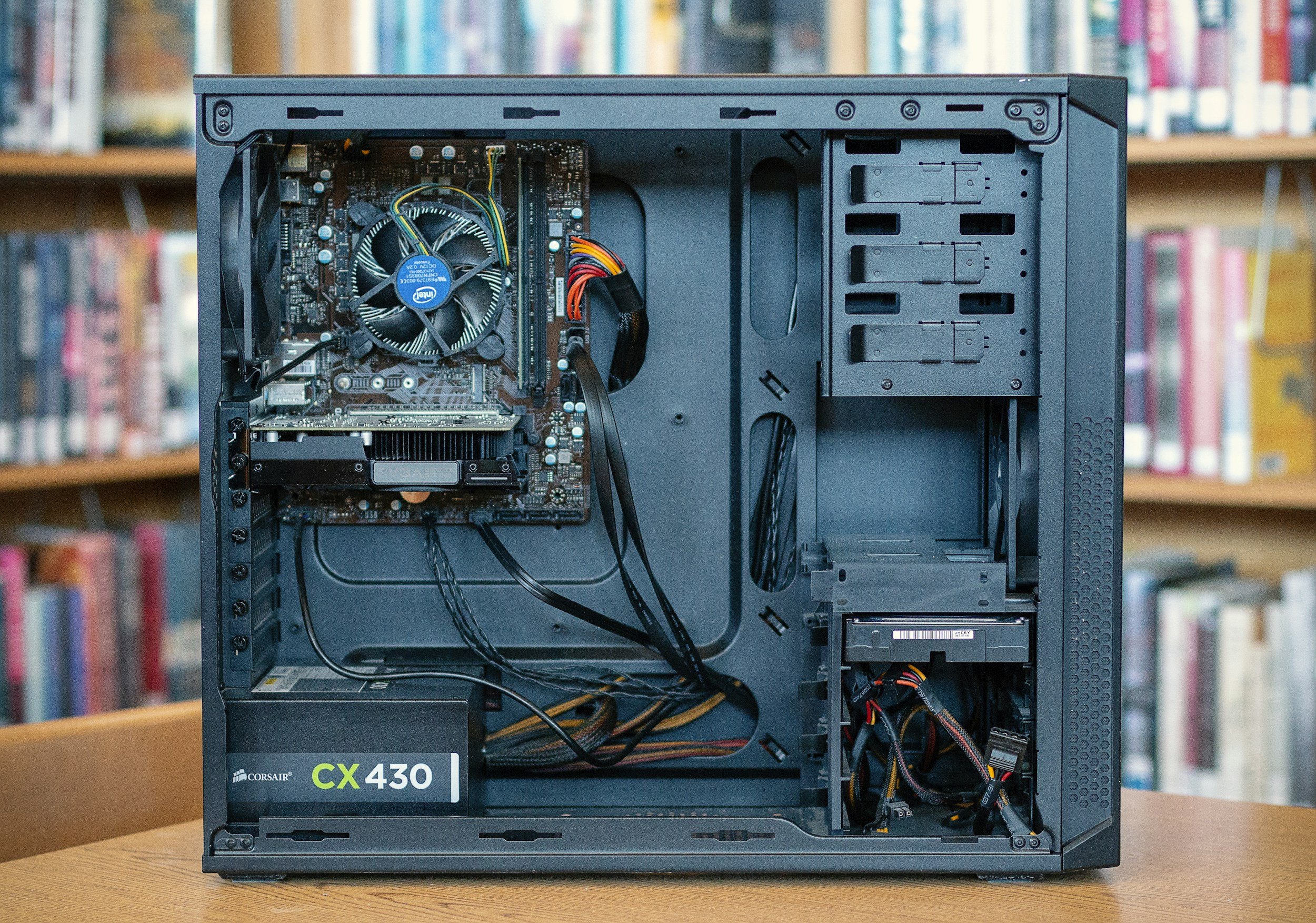 Open black computer case with internal components visible, including motherboard, cooling fan, and power supply, placed on a wooden table against a background of bookshelves.