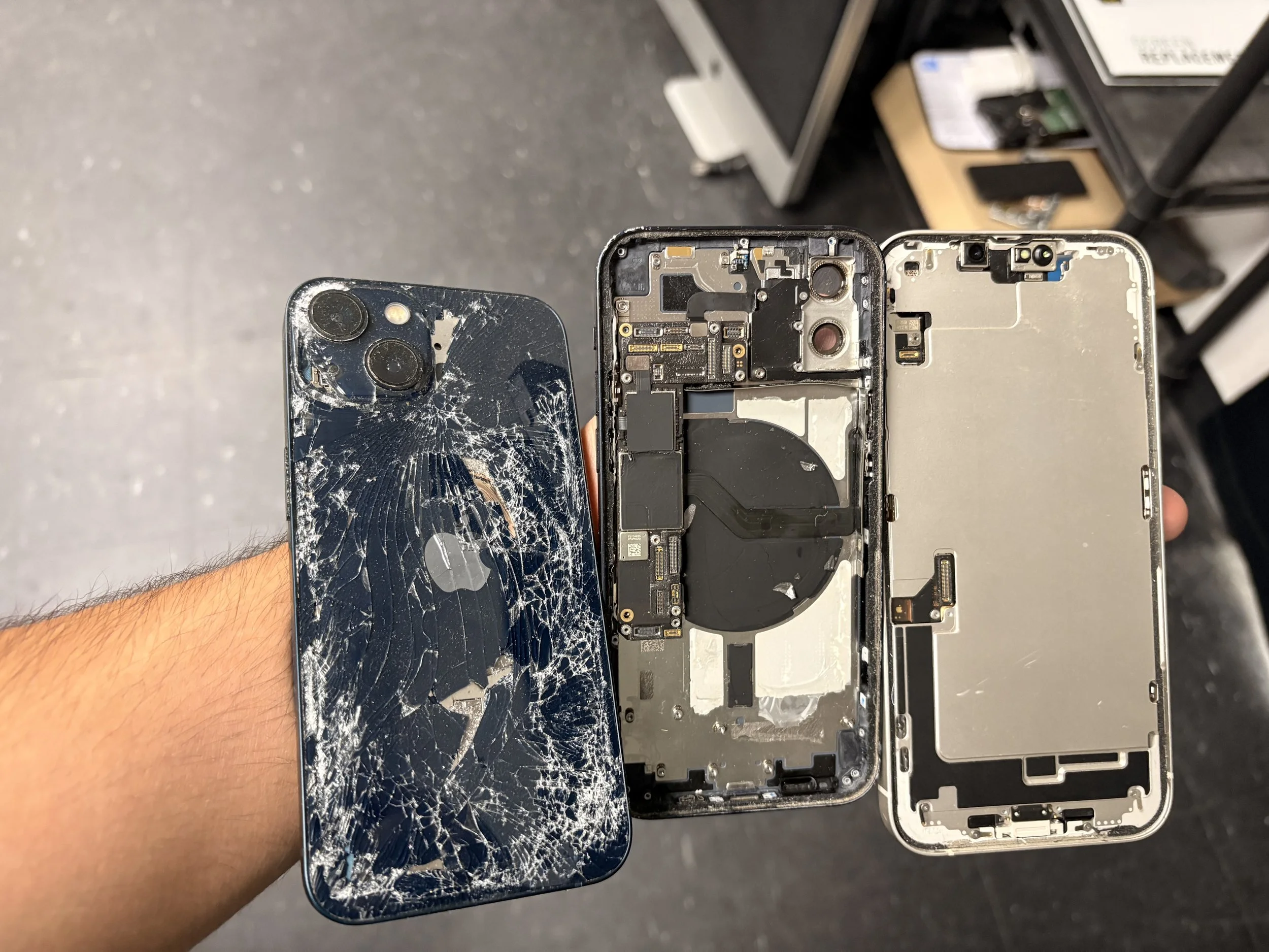A person holding a cracked iPhone with a shattered back glass panel, showing internal components, while the phone's screen is also damaged.