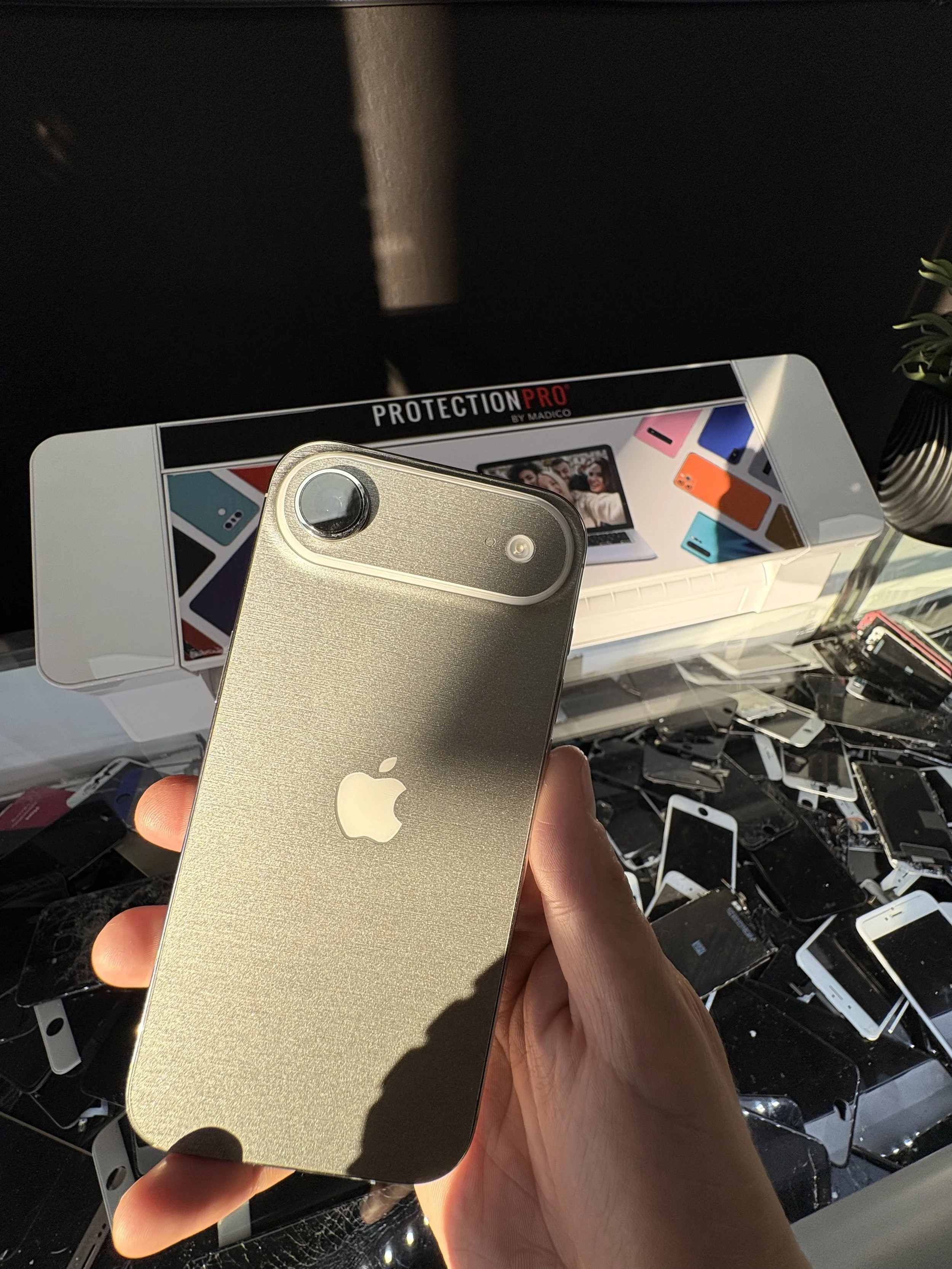 Hand holding a gold-colored iPhone with a circular camera lens on the top left corner, in front of a display box for ProtectionPro screen protectors with colorful phone images, surrounded by a variety of other phones and electronic devices on a store shelf.