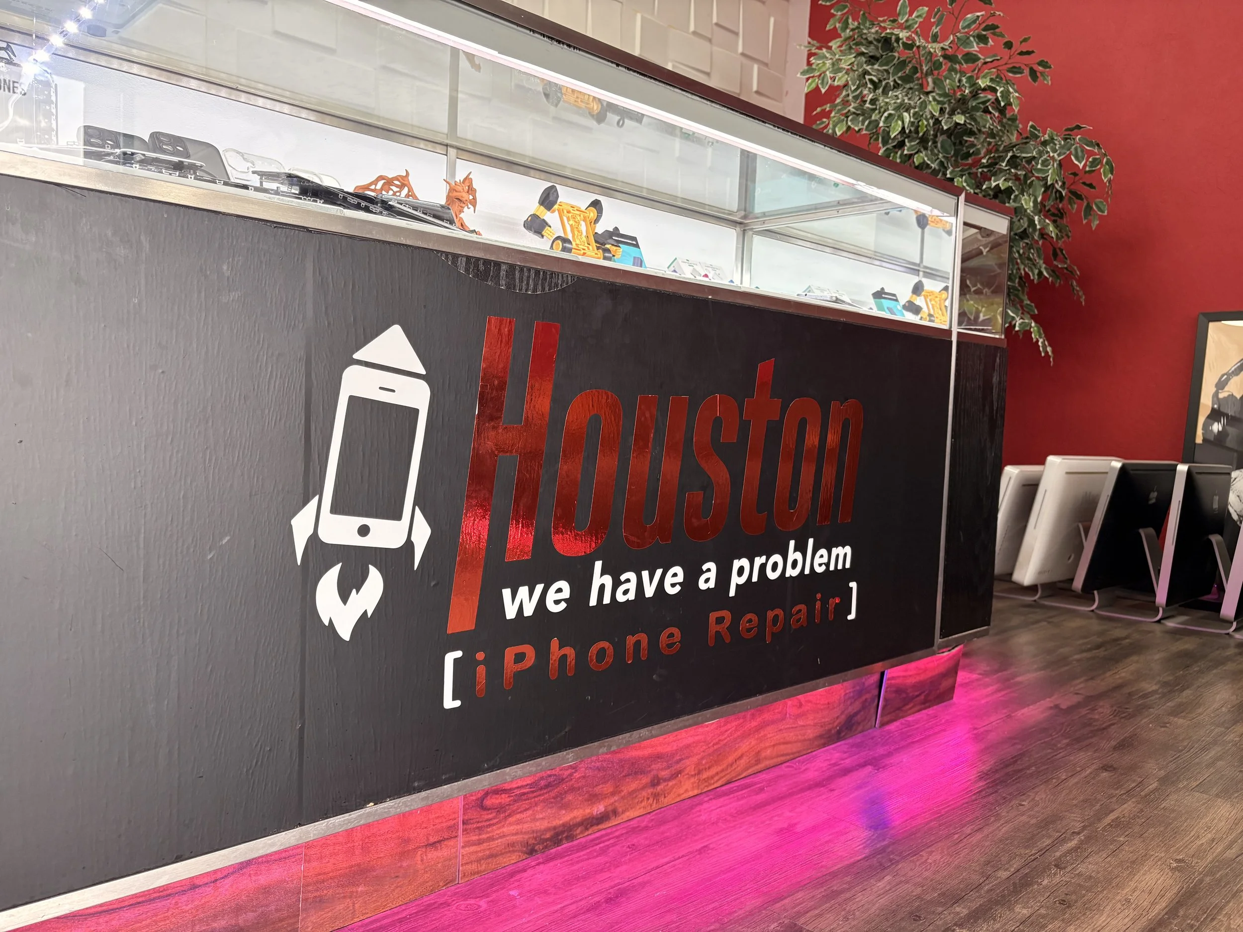 A black counter with a sign that says 'Houston' and 'we have a problem' with a graphic of a phone and a wrench, located inside an iPhone repair store, with a plant and red walls in the background.