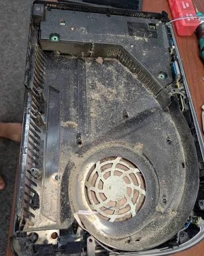 The inside of a dusty, partially disassembled gaming console with a large cooling fan and electronic components.