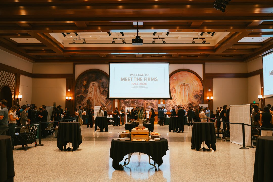 An indoor conference event hall filled with people standing and talking near tables. A large screen displays the text "Welcome to Meet the Firms Fall 2024." The room has decorative murals on the walls and tables with food in the foreground.