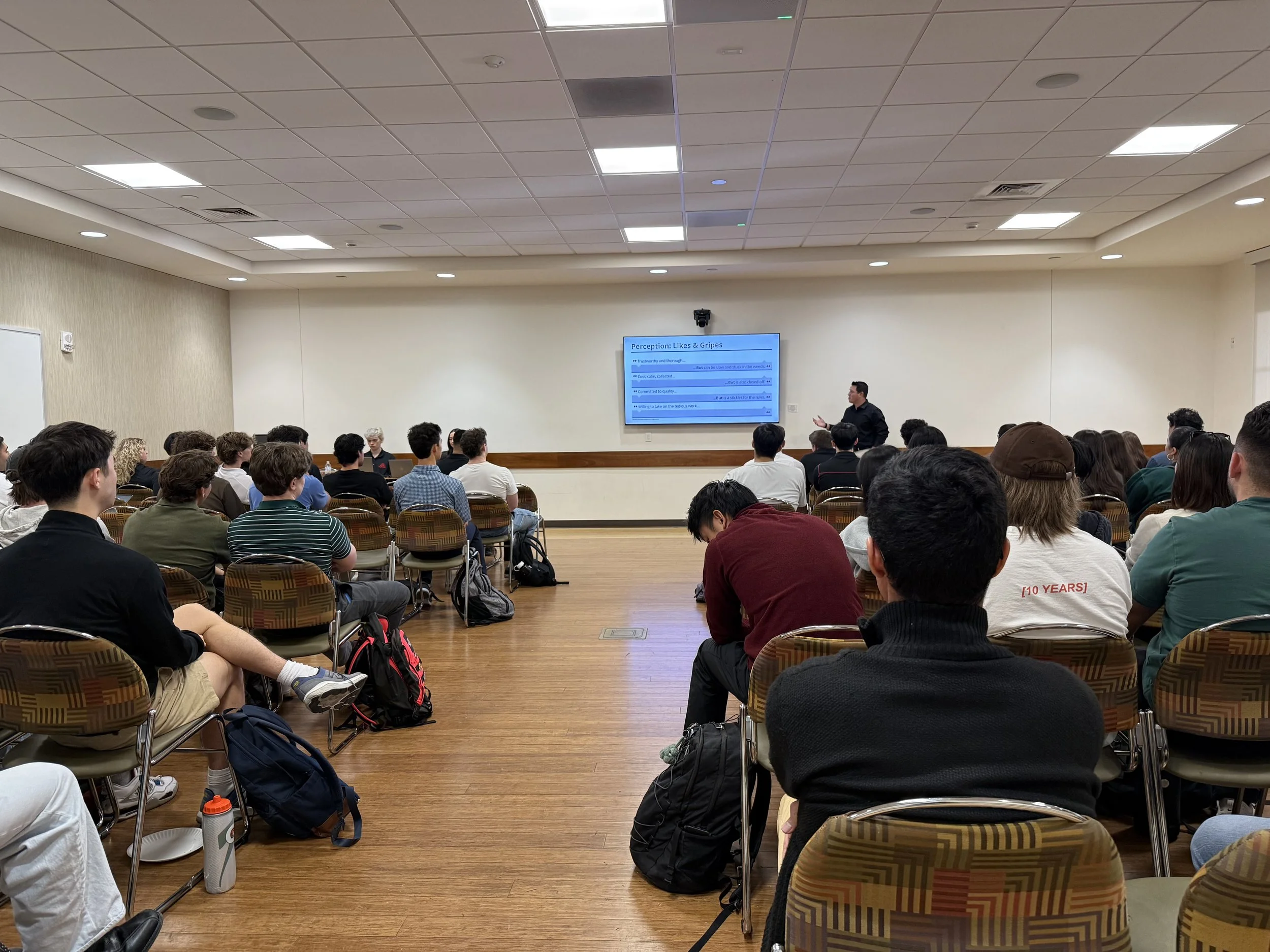 A classroom or conference room filled with students and a speaker at the front. The audience is attentively listening, and a large screen displays a presentation titled 'Perception: Likes & Gripes.' The room has a white ceiling with square lights, light-colored walls, and a wooden floor. Students are seated in chairs with backpack and water bottles nearby.