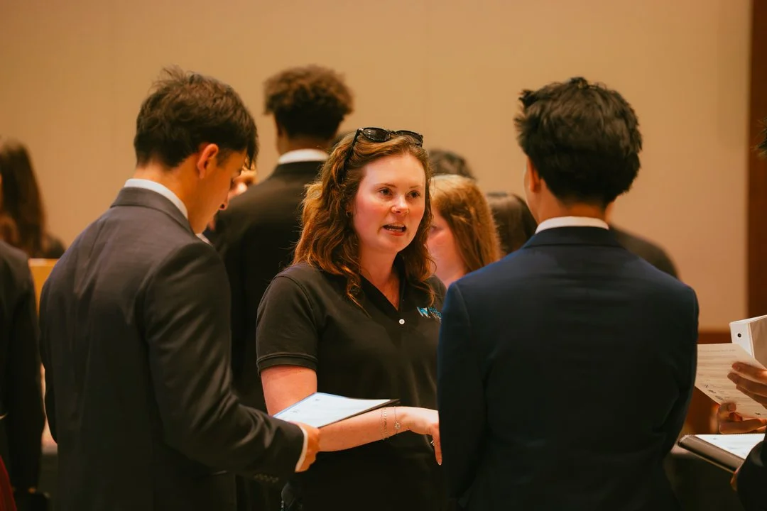 Woman in black shirt talking to two young men in suits at a professional event.