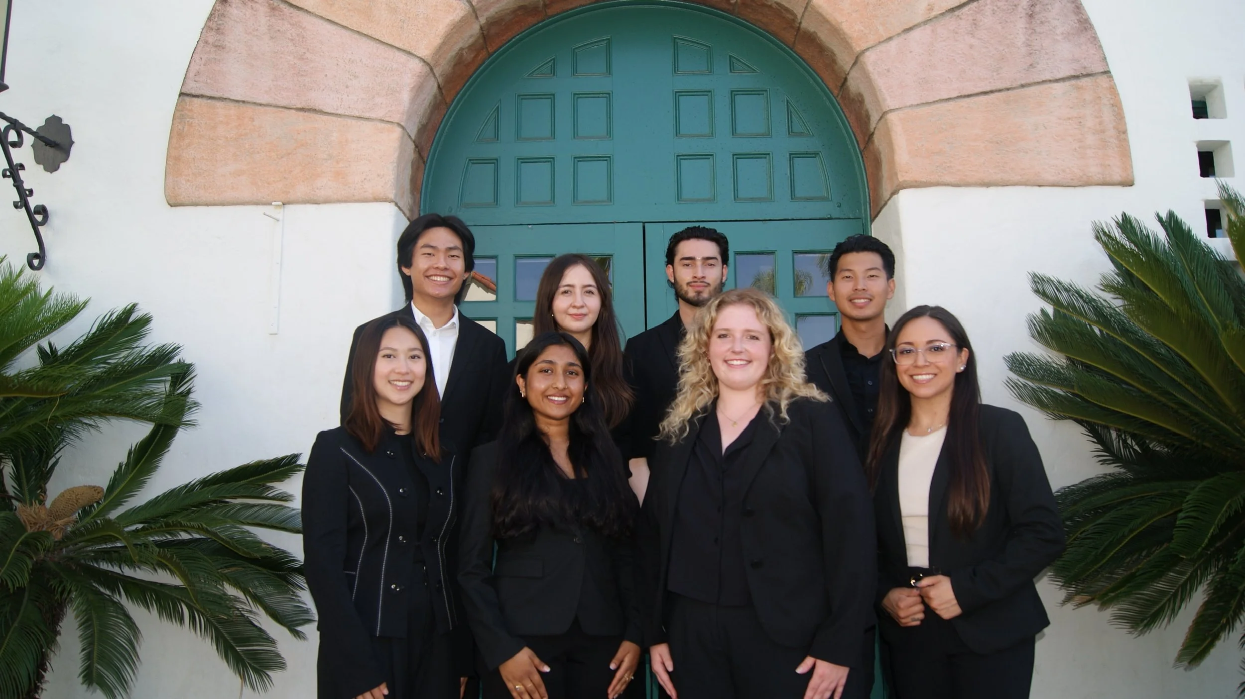 Group of eight young professionals, five women and three men, standing outdoors in front of a large green door with an arch, surrounded by palm plants, smiling at the camera.