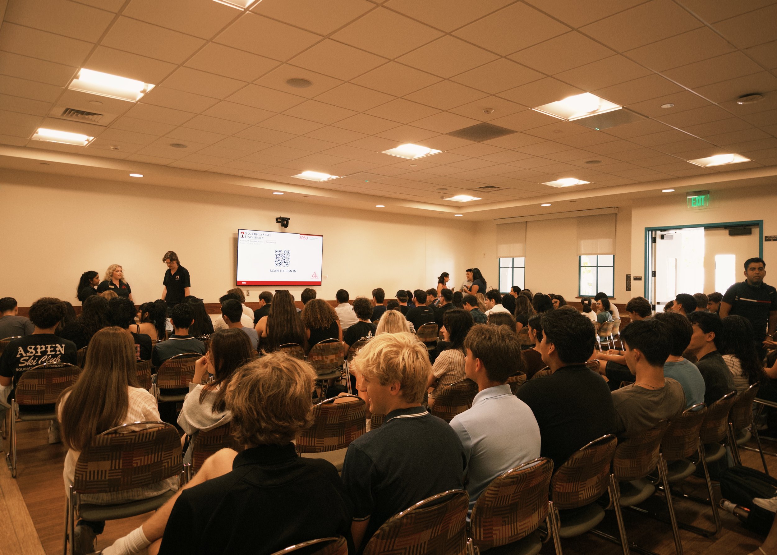 People attending a lecture or conference in a large room with a projector screen and many chairs.