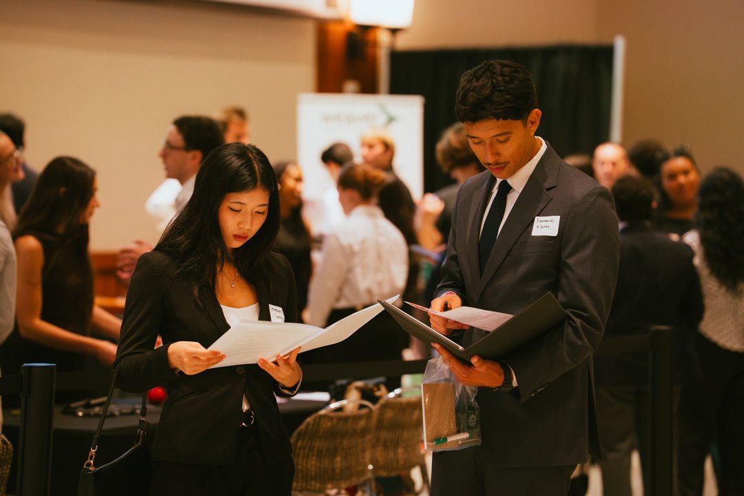 Two young professionals, a woman and a man, dressed in business attire, are looking at documents at a networking event or conference with many other people in the background.