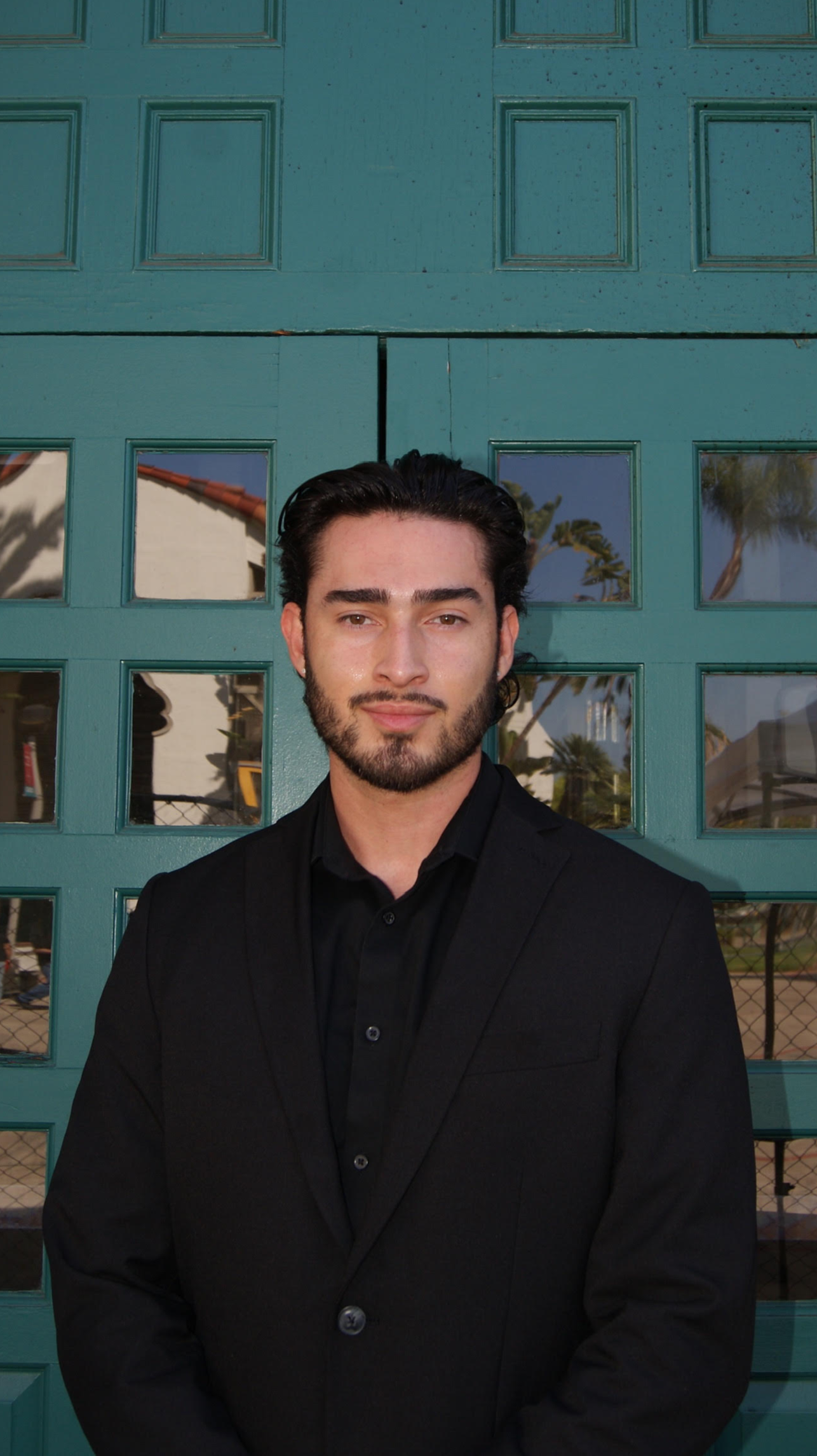 A man with dark hair, light skin, and facial hair wearing a black suit and black shirt, standing in front of a teal double door with glass panels reflecting outdoor scenery, in a sunny environment.