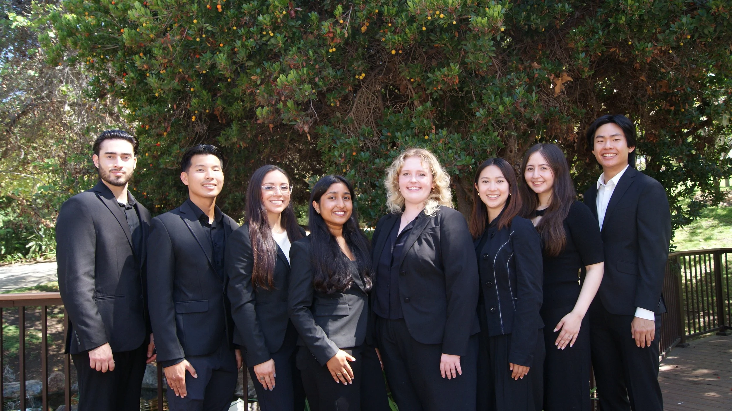 Group of eight diverse young adults dressed in formal black suits standing outdoors in front of green trees, smiling at the camera.