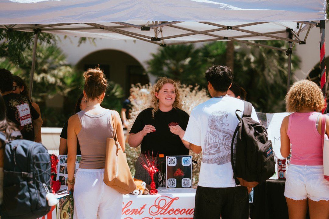 A young woman with curly blonde hair talks to a man at an outdoor event booth, with other people standing around and palm trees in the background.