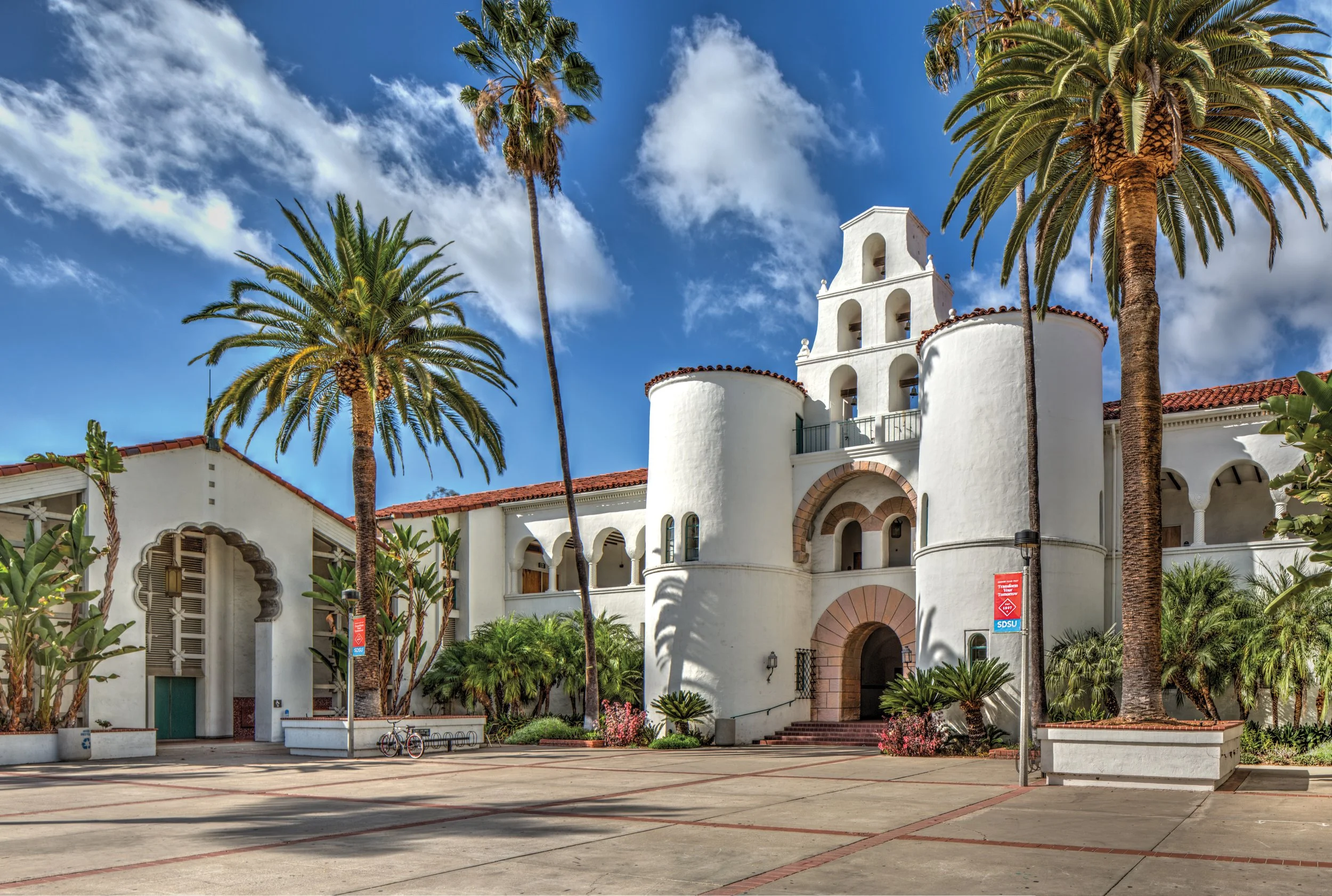 A white Mediterranean-style building with arched doorways and windows surrounded by tall palm trees and landscaped greenery under a partly cloudy blue sky.