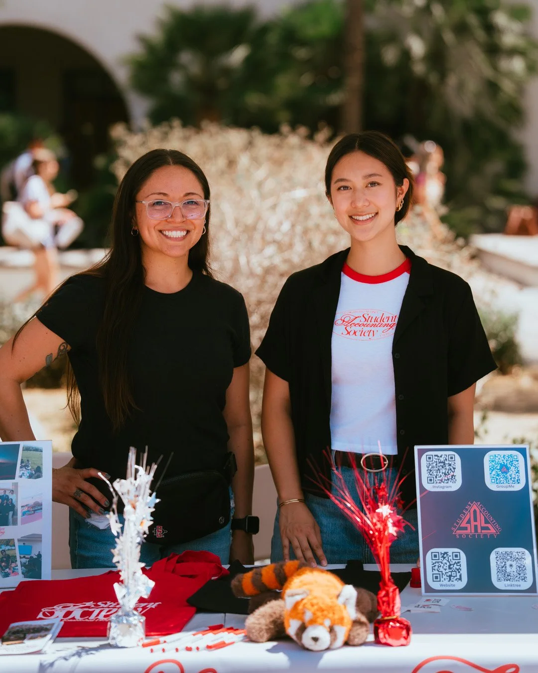 Two women smiling at a booth with promotional materials and stuffed animal on a table outdoors.
