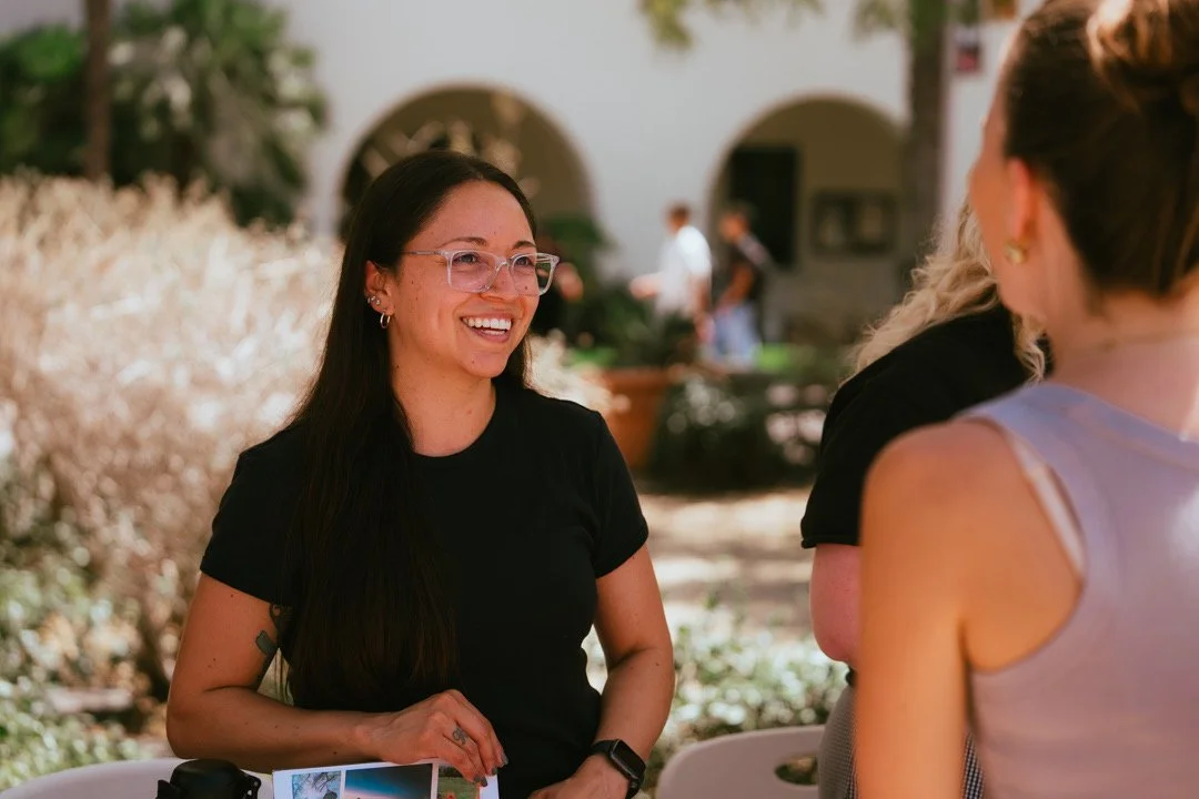 A woman with long dark hair, glasses, and a black shirt smiling and talking to two women outdoors.