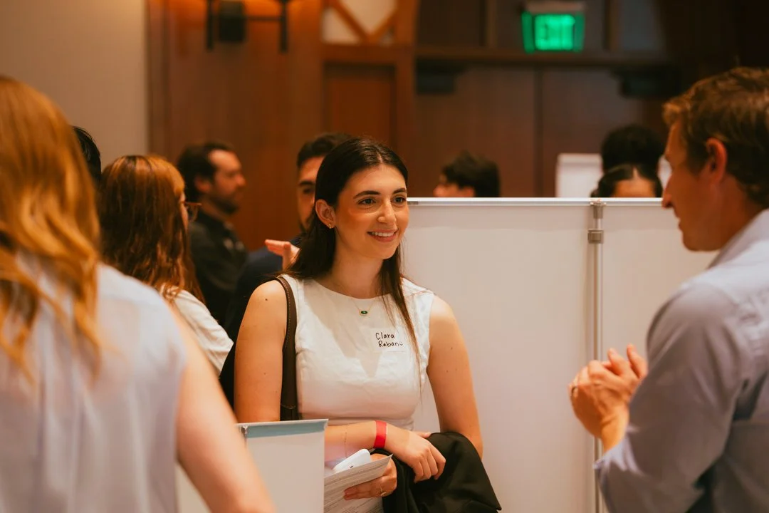 A woman with long dark hair, wearing a white sleeveless dress, smiling and talking to a man at a professional event or conference, with other attendees in the background.