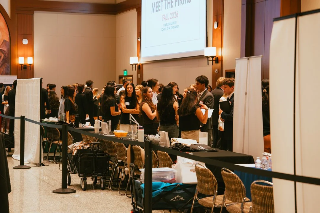 A large group of people in formal attire gathered in a conference room, engaging in conversations. A large screen displays information about a fall 2026 event, and there are tables with materials and water bottles.
