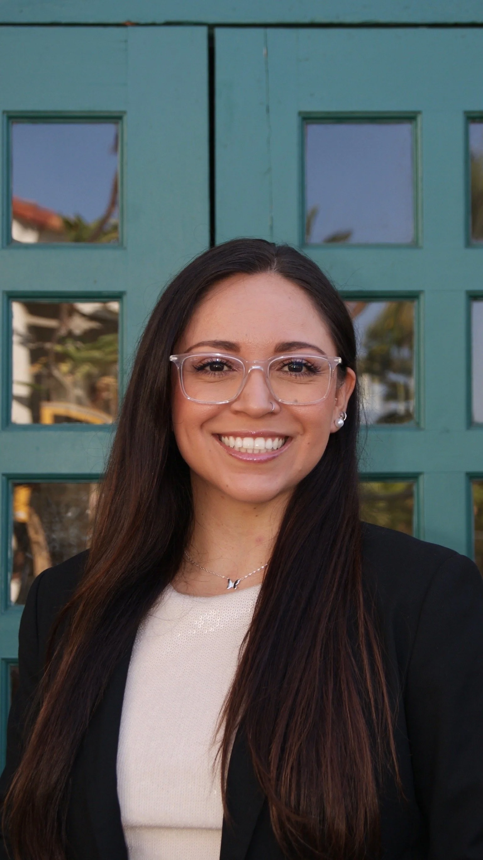 Young woman with long dark hair, wearing glasses, a pearl earring, a necklace, a black blazer, smiling, standing in front of a teal-colored door with square glass panels.