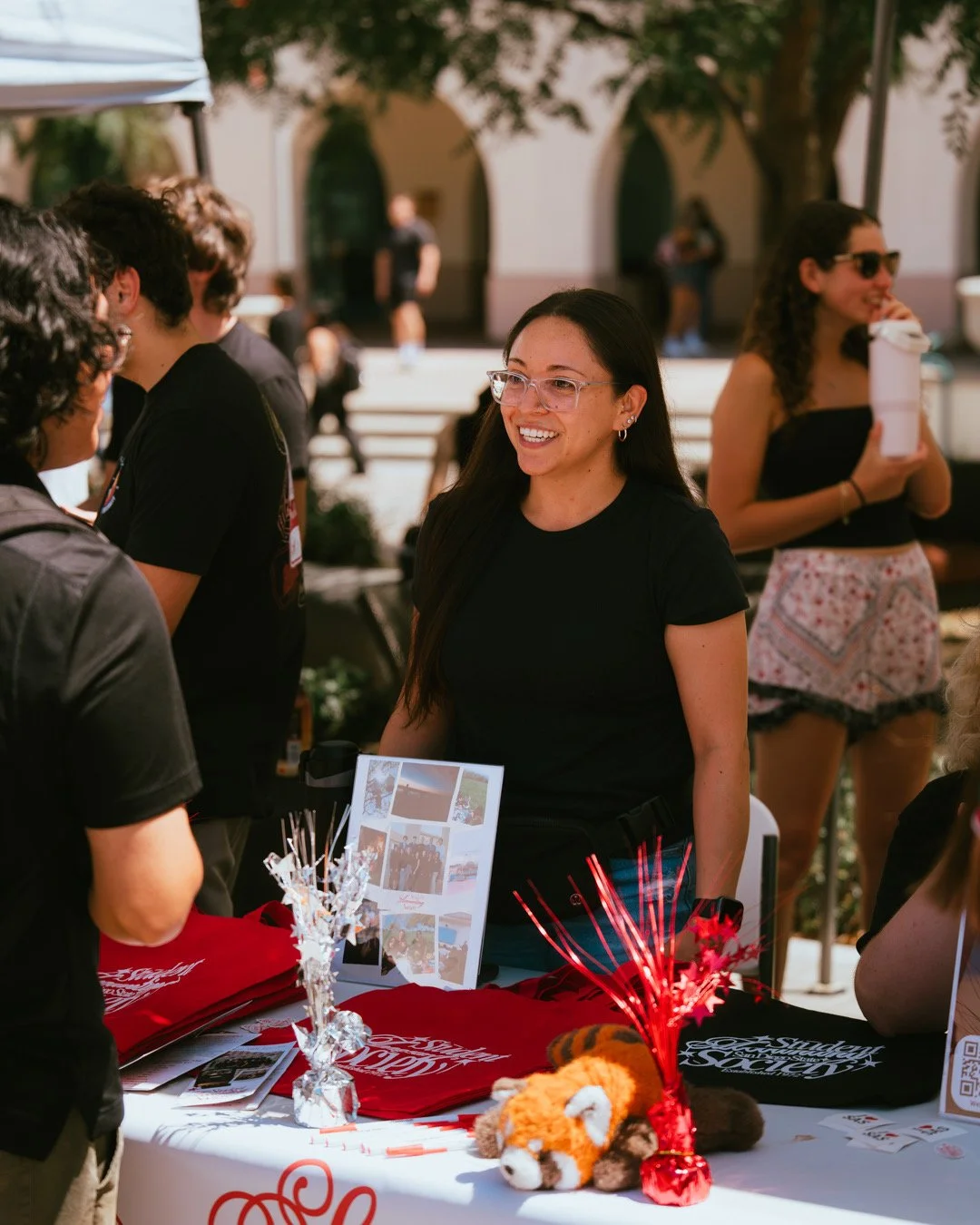 A woman with glasses smiling and talking to a person at an outdoor booth during daytime. The booth has red and black T-shirts, a plush tiger toy, and holiday decorations.