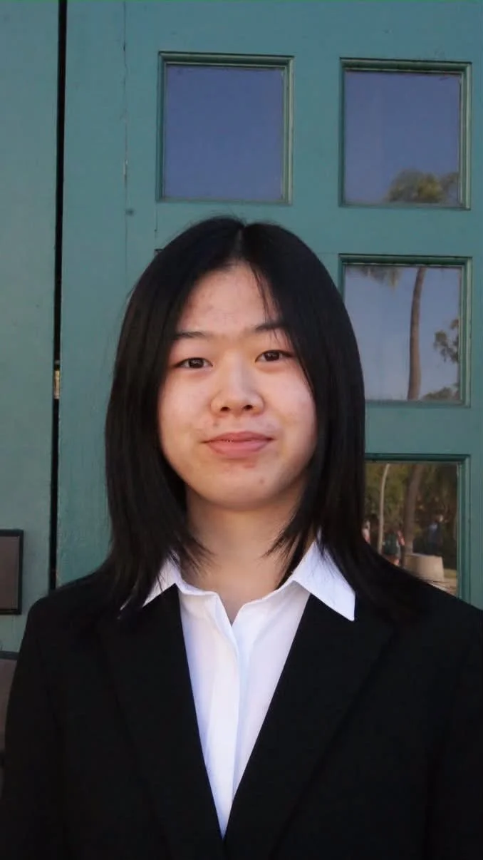 Young woman with shoulder-length black hair, wearing a white collared shirt and black blazer, standing in front of a green door with square windows.