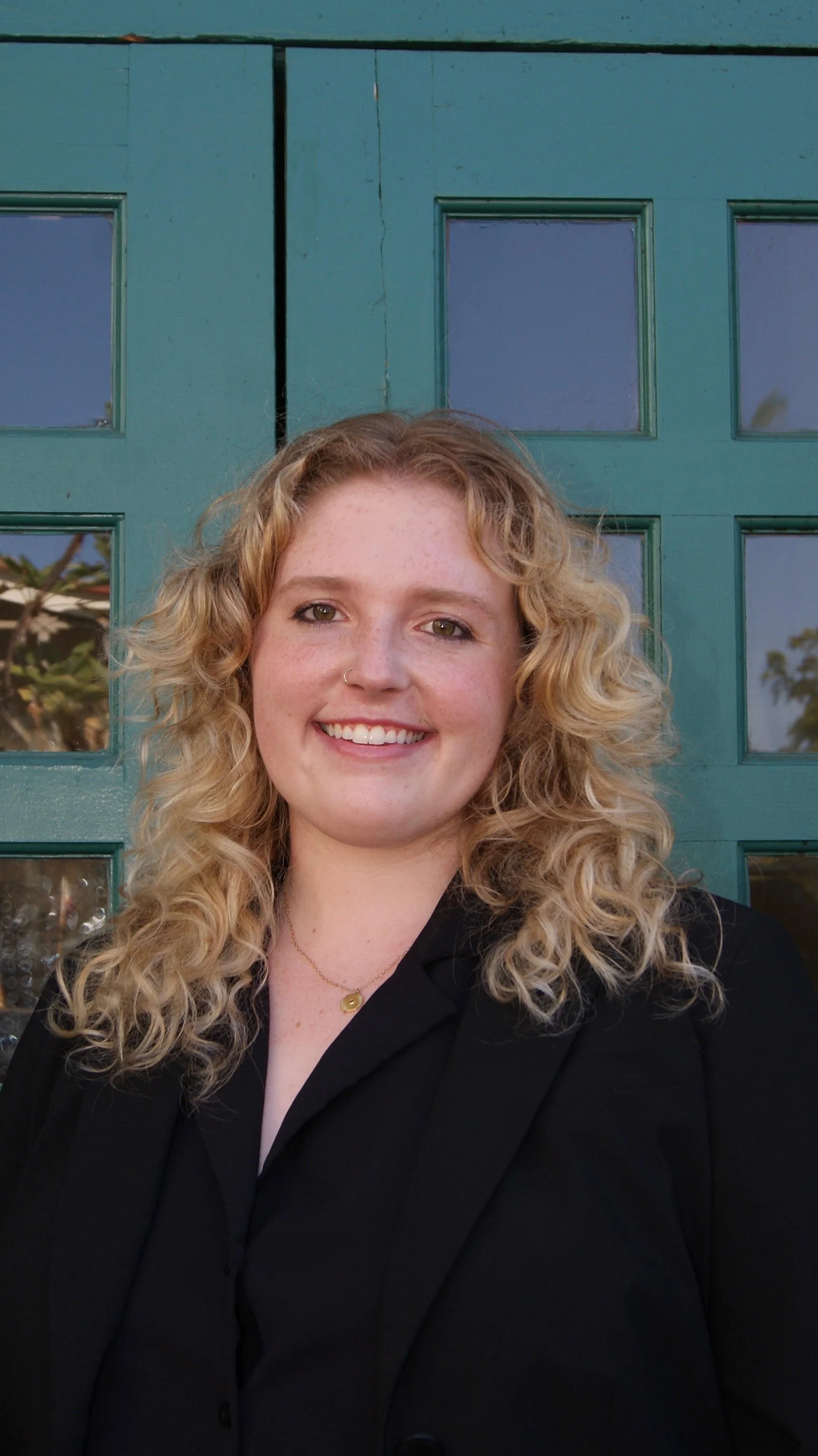 Portrait of a young woman with curly blonde hair, smiling, wearing a black blazer, standing in front of green double doors with glass panes.