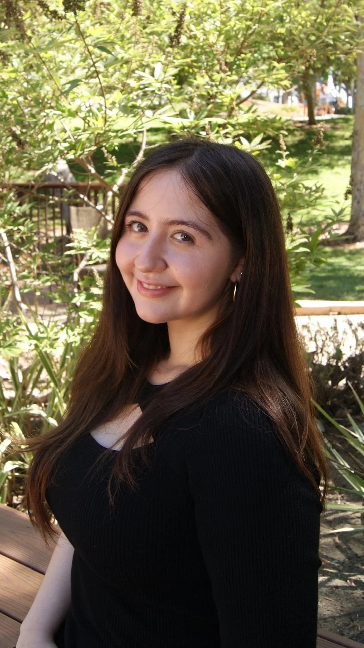 A young woman with long brown hair smiling outdoors, with green trees and a wooden bench in the background.
