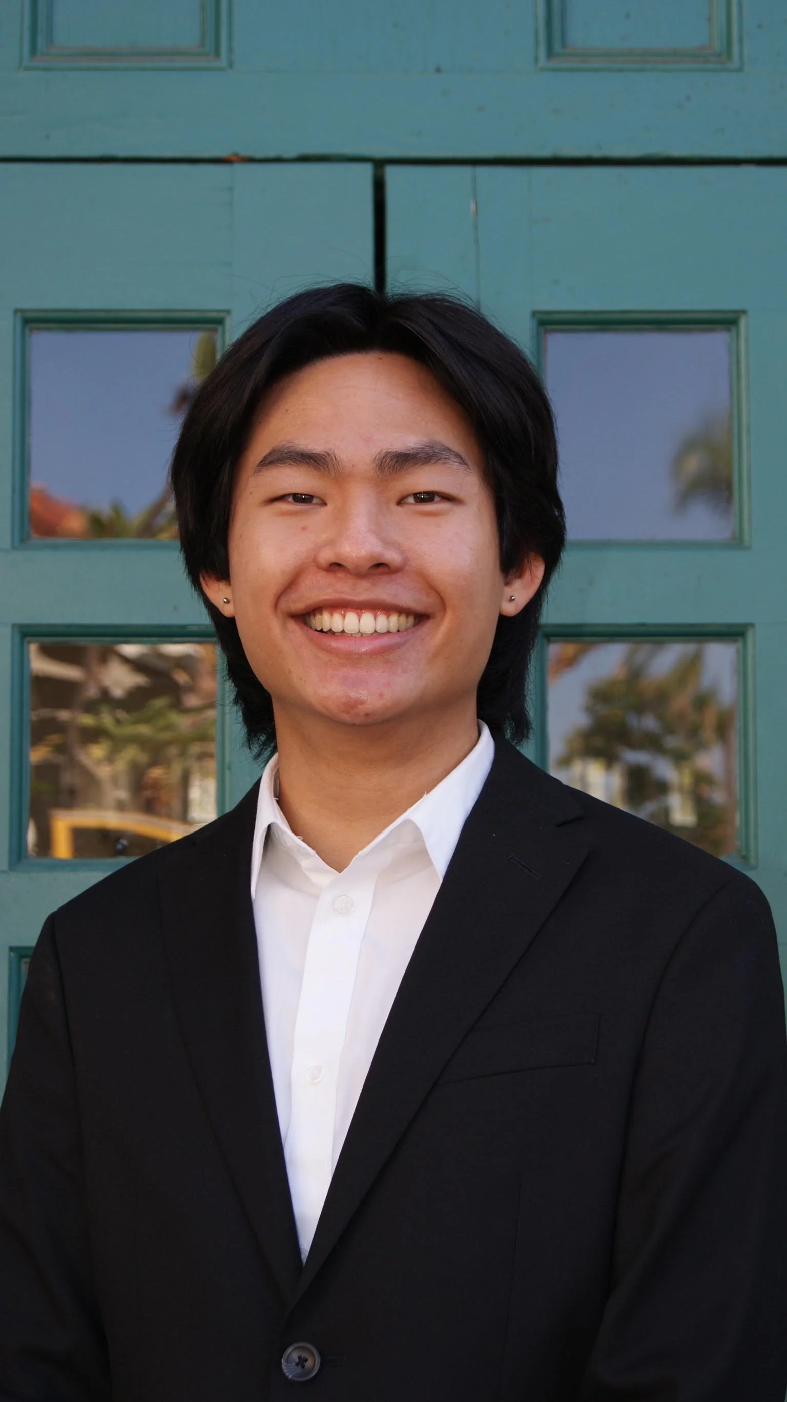 A young Asian man with black hair, smiling, wearing a white shirt and black suit jacket, standing in front of a green door with glass panels.