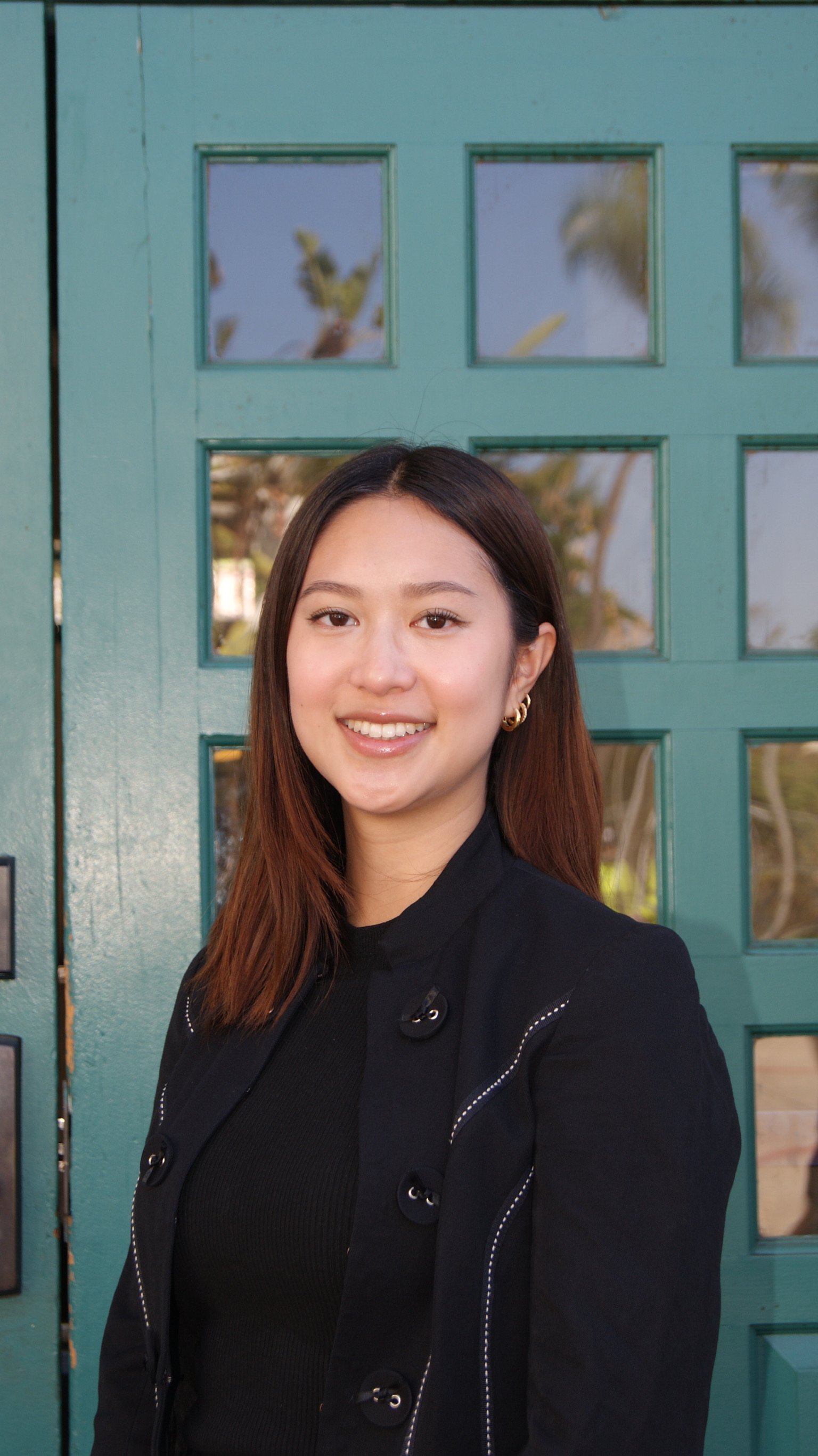 A young woman with long brown hair, wearing a black top and a black jacket with metal buttons, smiling in front of a teal door with square glass panes, some of which reflect trees and the sky.