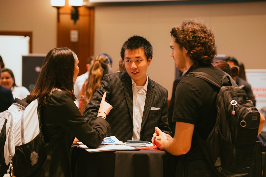Three people engaged in conversation at a conference or networking event, with a group of people blurred in the background.
